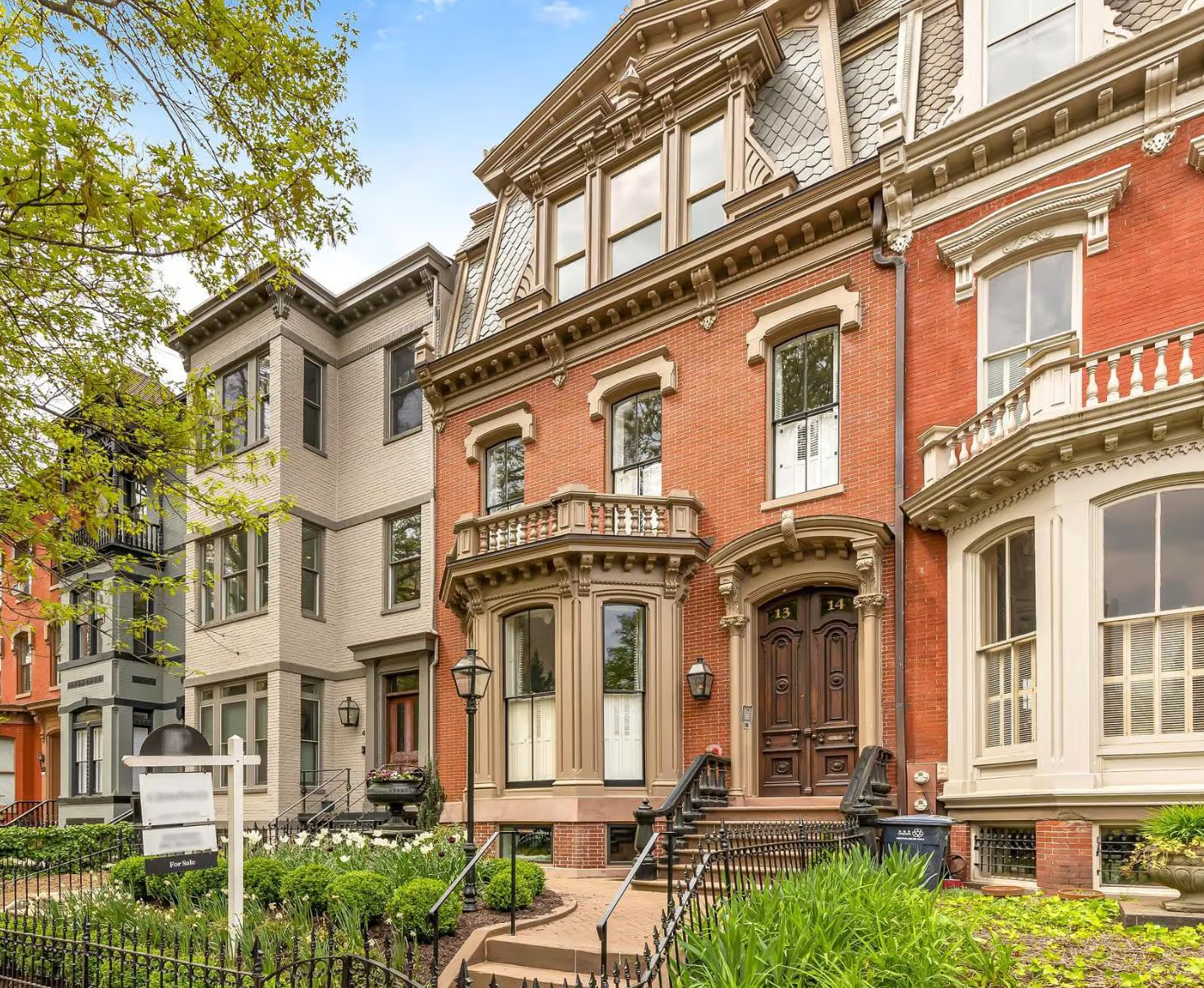 Row of historic brick and stone houses with ornate facades, front gardens, and steps leading to their entryways.