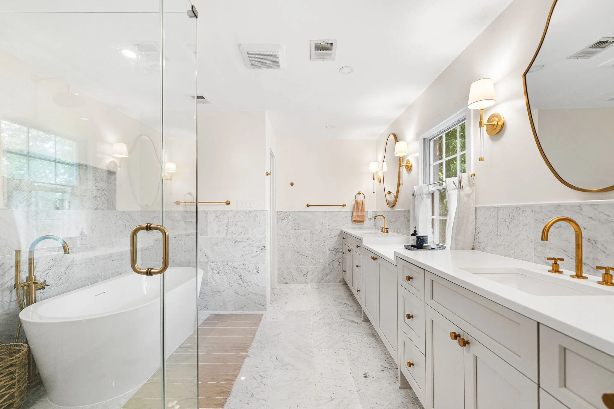 A bright, modern bathroom with white cabinetry, gold fixtures, and a large mirror. There is a glass-enclosed shower, a freestanding bathtub, and a window with white curtains. The decor features minimalistic, elegant design with white and gold accents.