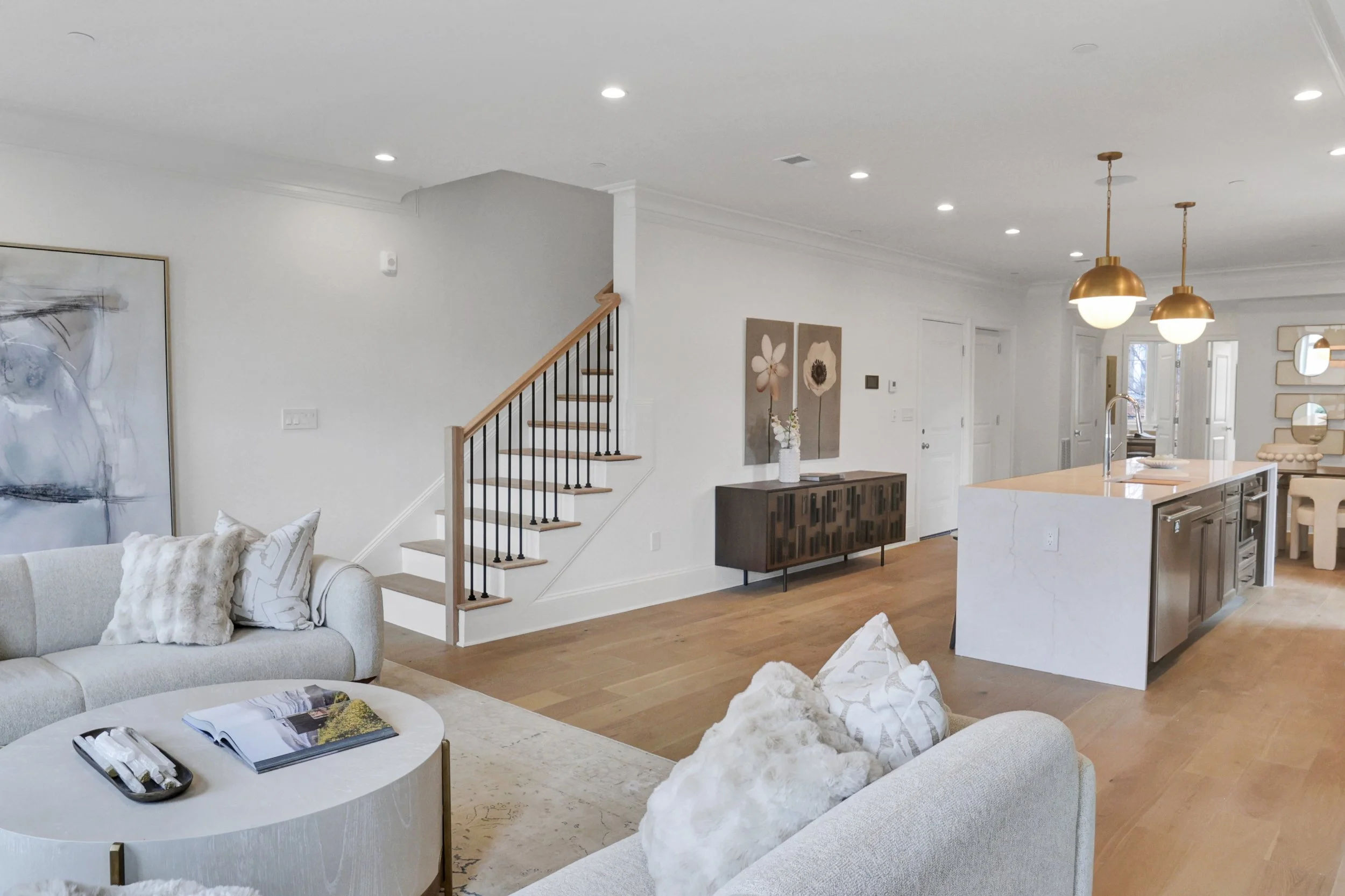 Living room with white couches, pillows, coffee table with magazine, abstract art, staircase, wooden floors, pendant lights, and open kitchen in the background.