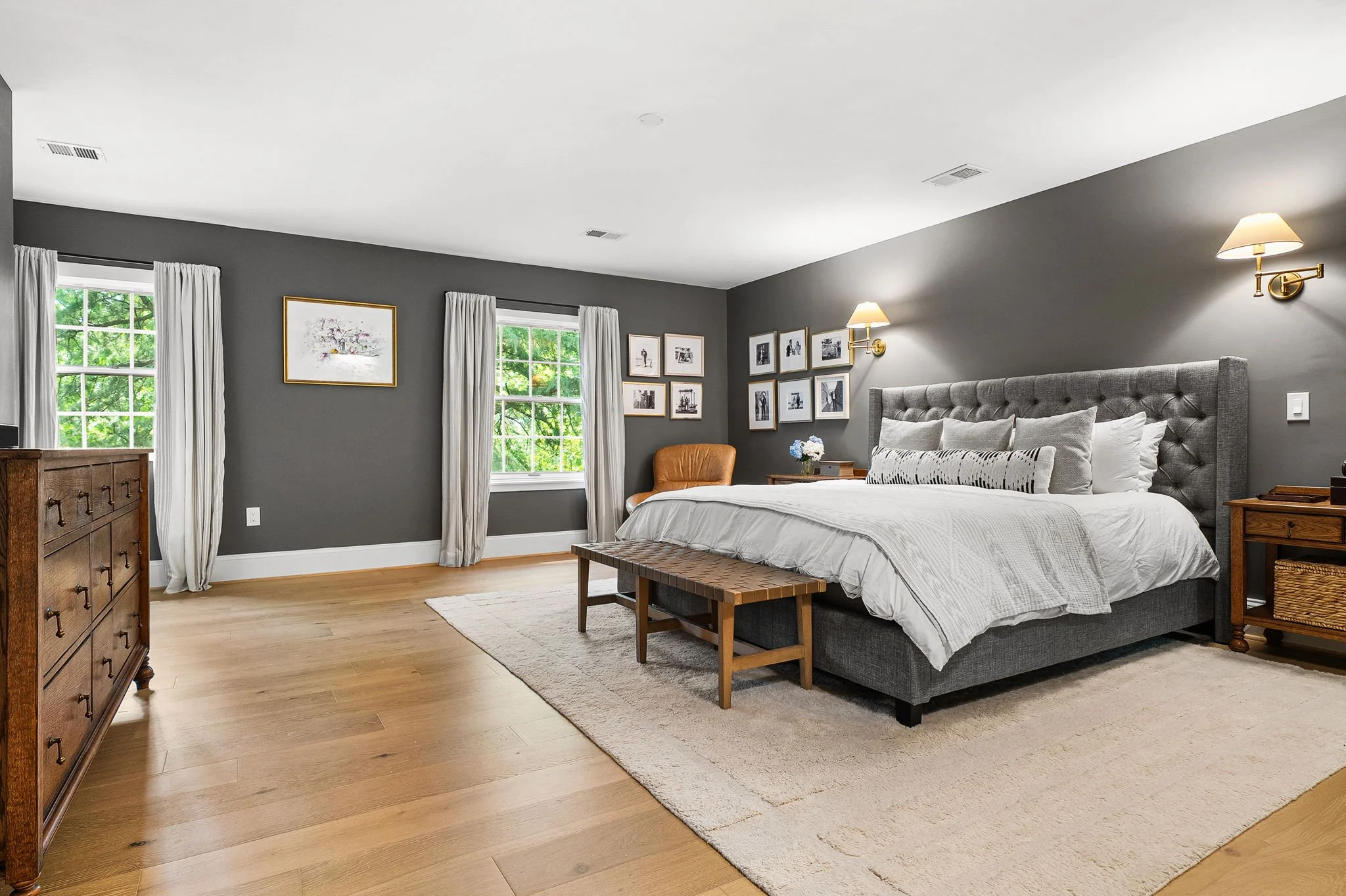 A bedroom with dark gray walls, two windows with white curtains, a bed with gray upholstered headboard, white bedding, and framed pictures on the wall. There are two wall lamps on either side of the bed, a wooden dresser, and a small wooden bench at the foot of the bed. The floor is light wood with a beige area rug.