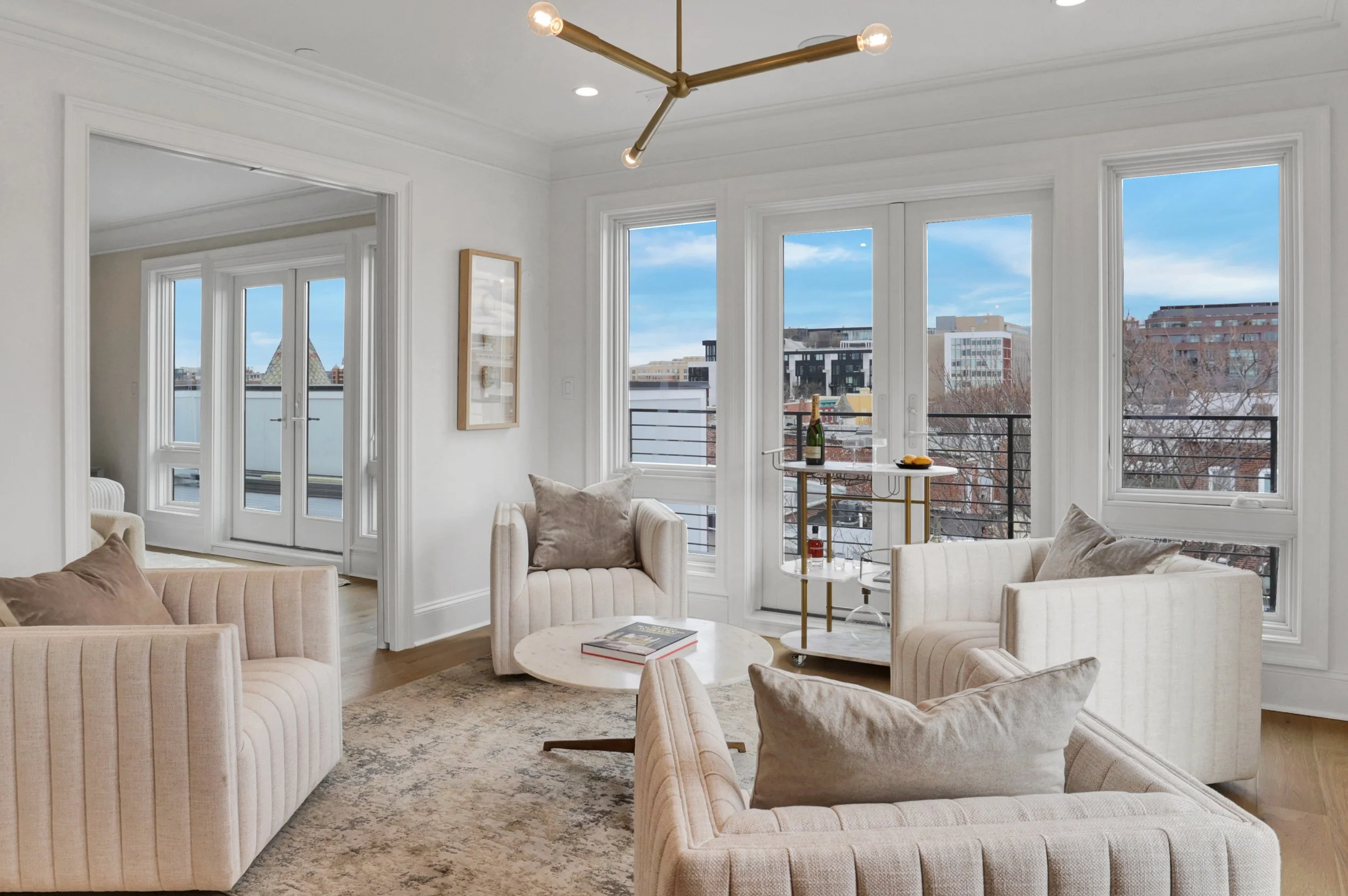 Bright living room with multiple large windows showing city buildings and blue sky, beige tufted sofas, a round coffee table with books, a gold chandelier, and a small bar cart with a bottle of champagne and glassware.