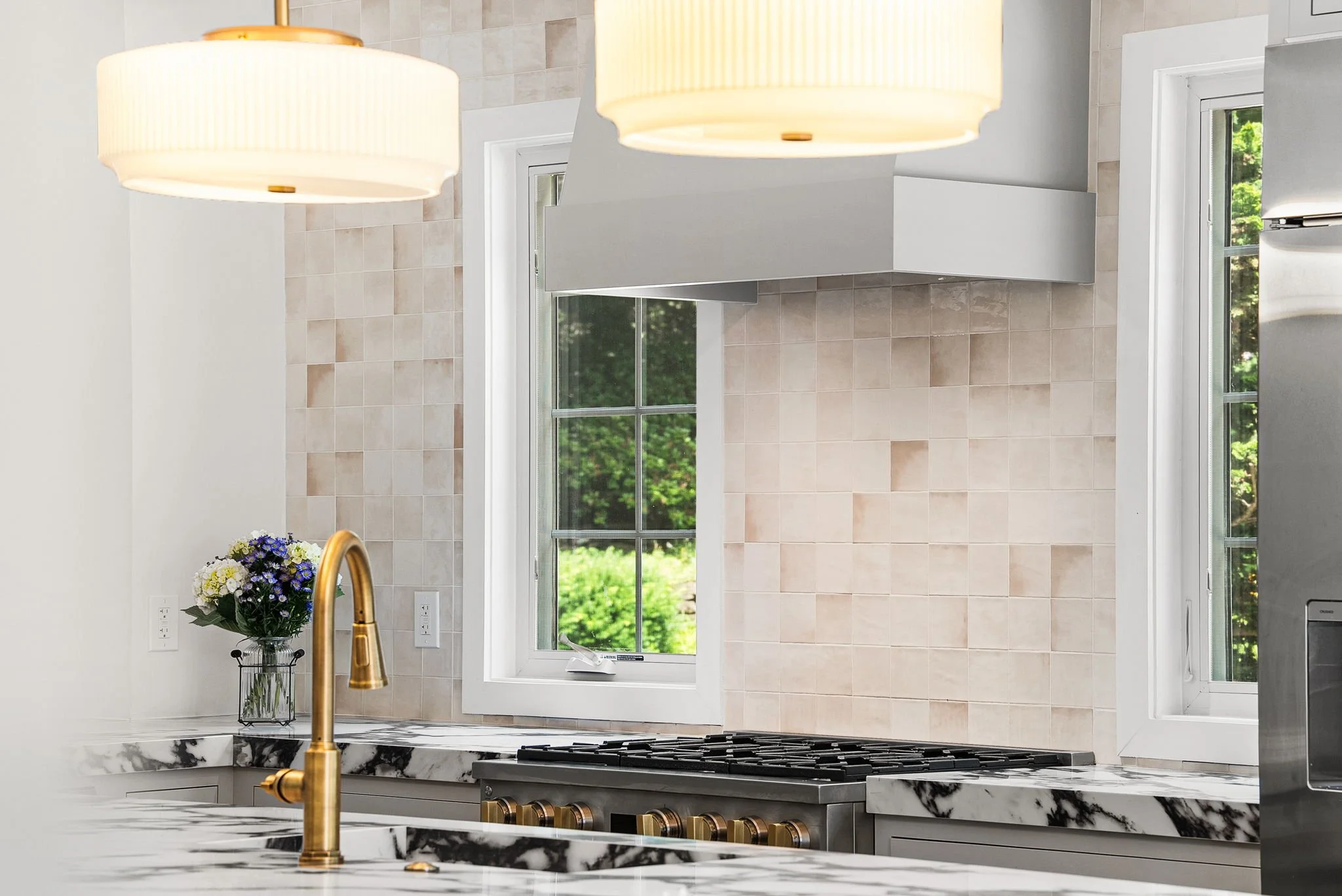 Modern kitchen with white cabinets, a marble countertop, and a black stove. There are large windows with views of green foliage, and a bouquet of purple and white flowers on the counter. Two large pendant lights hang from the ceiling.