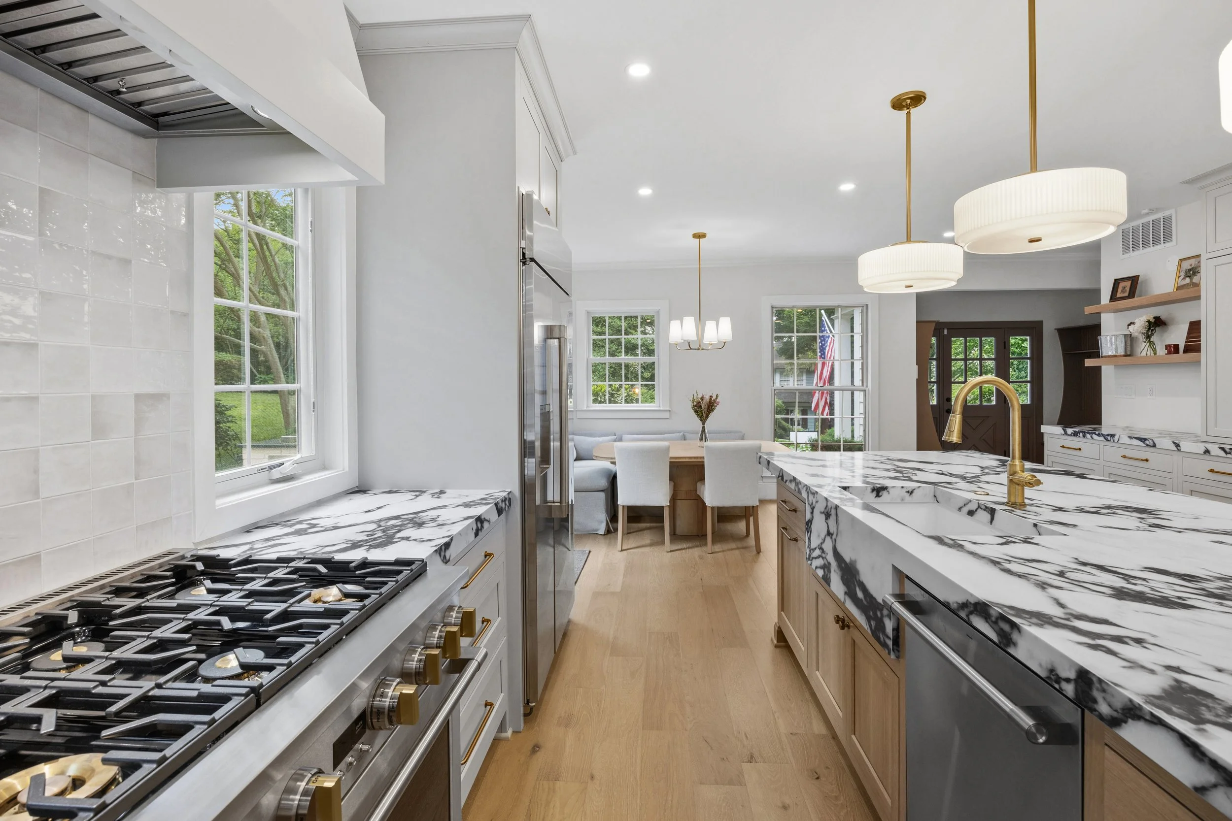Modern kitchen with white cabinets, marble countertops, gold fixtures, and large windows. A dining area with a white table and chairs is visible in the background.