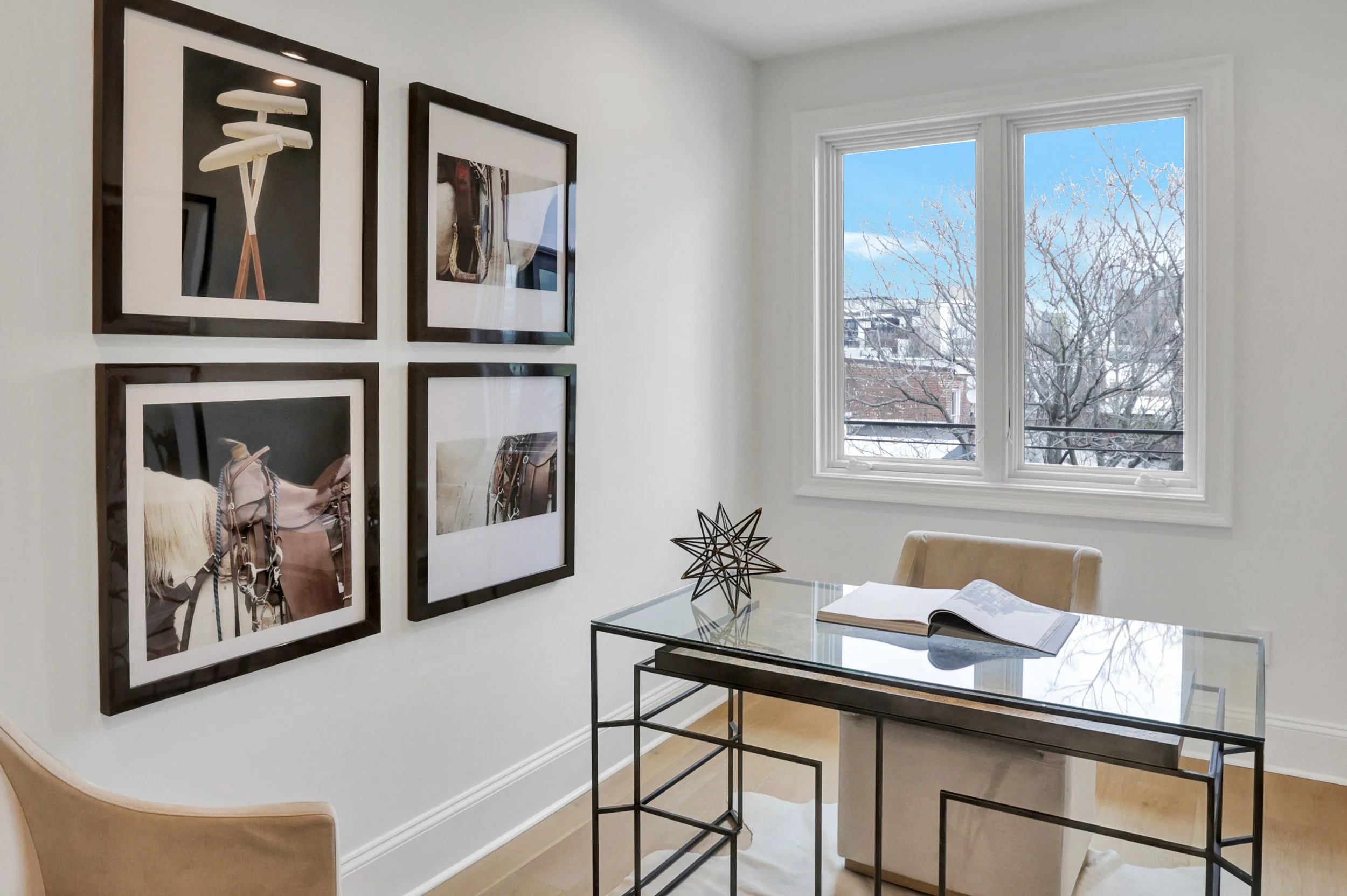 Interior of a modern room with white walls, large window showing a tree and buildings outside, wall art of four framed photographs, glass table with a decorative star sculpture, open book, and beige chairs.