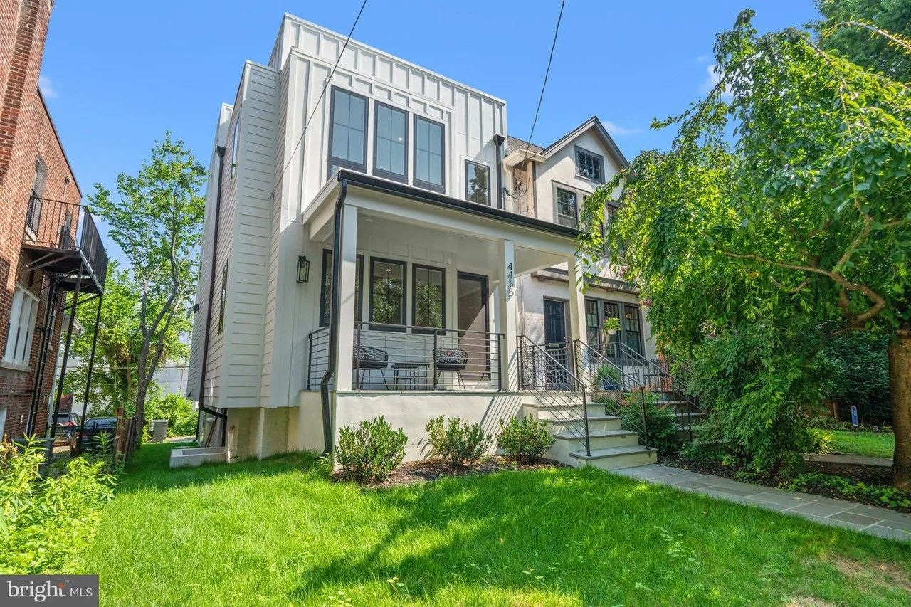 Front view of a modern, multi-story residential house with a small porch, metal railing, and stairs leading to the entrance. The house has white siding and large windows, with a lush green lawn and trees surrounding it.