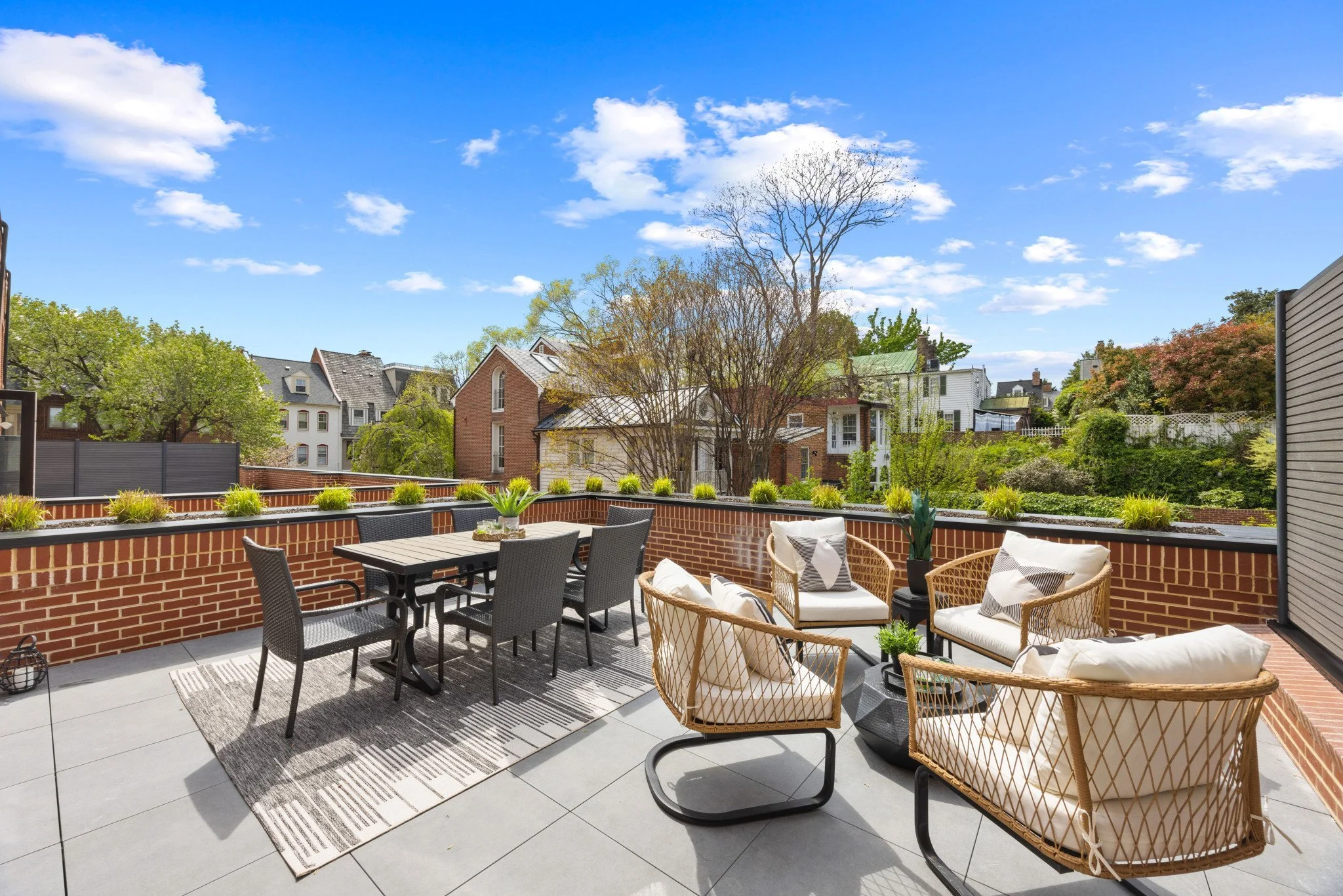 Outdoor patio with black and wicker chairs, a table, and cushions on wicker chairs, enclosed by a brick wall with potted plants, overlooking trees and neighboring houses with blue sky and scattered clouds.