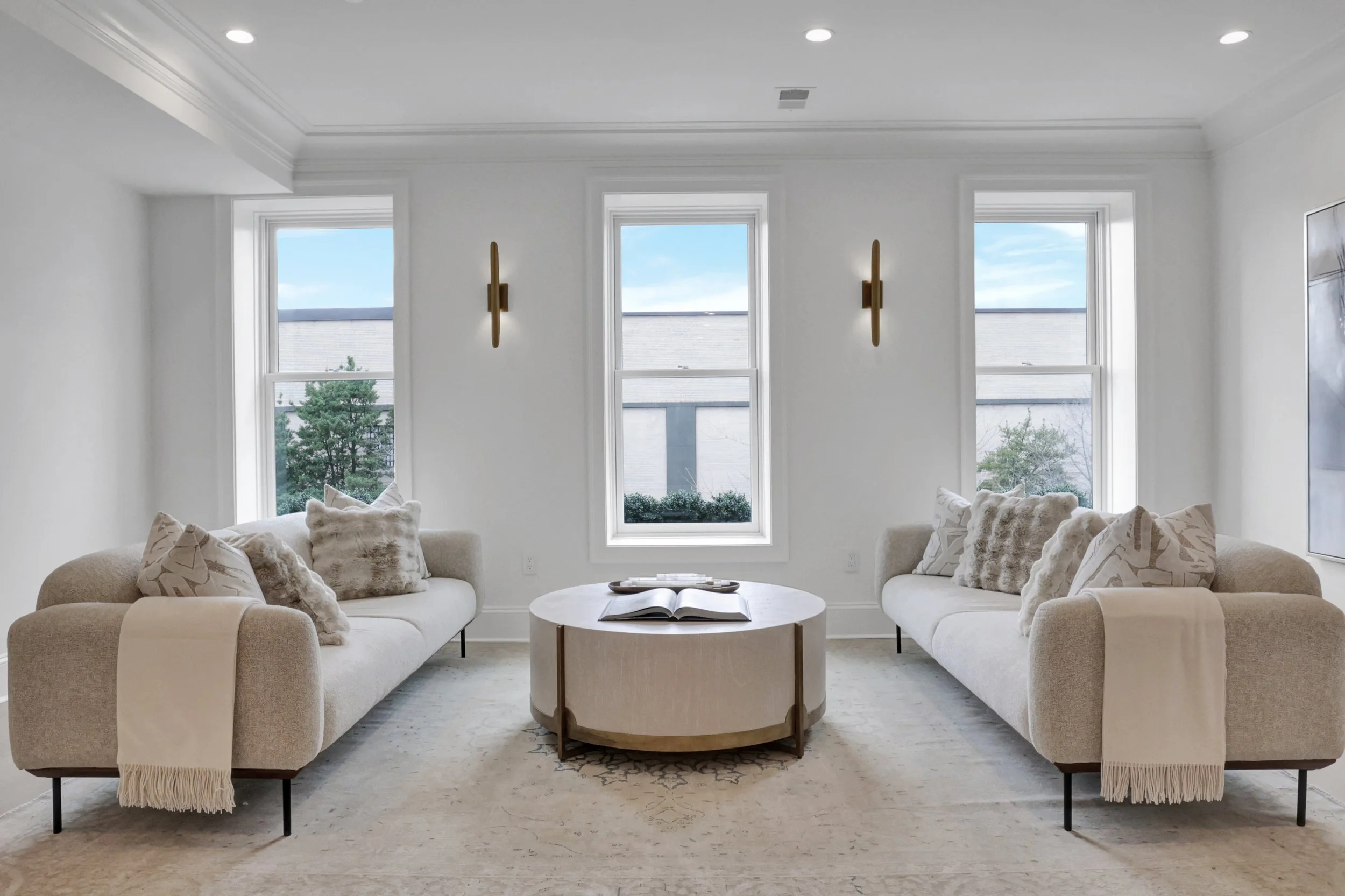 A living room with three large windows, two beige sofas with pillows, a round coffee table with an open book, and modern wall sconces.