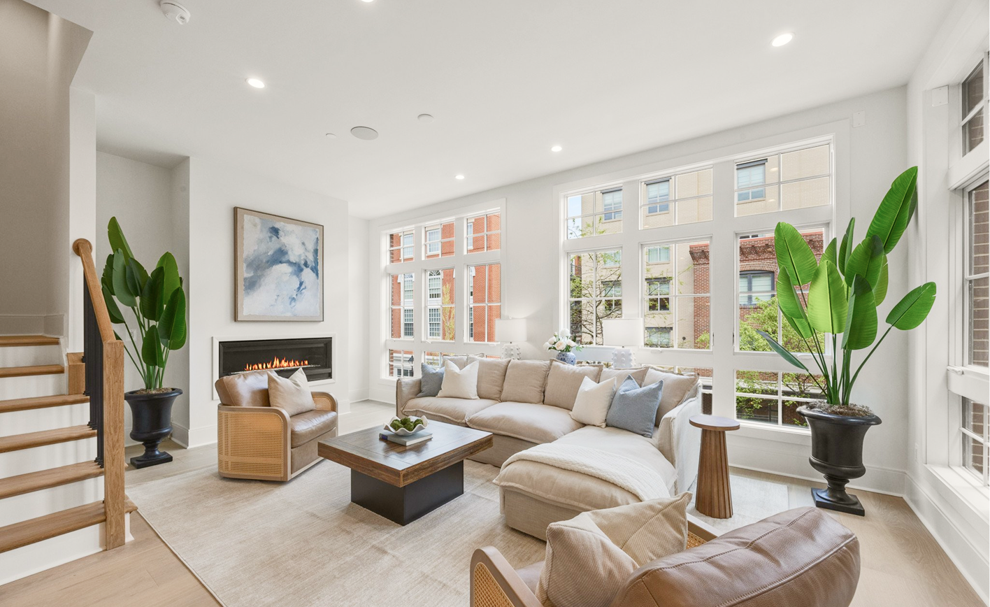 Bright living room with large windows, beige sectional sofa with white and blue pillows, wooden coffee table, wall-mounted fireplace, abstract art above fireplace, house plants in black pots, light-colored wooden flooring, staircase with wooden steps on the left.