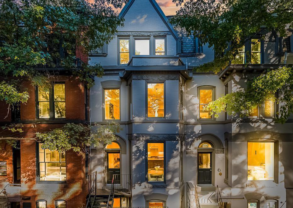 Three-story Victorian-style row house with a white facade, black door, and illuminated windows at sunset, partially obscured by trees.