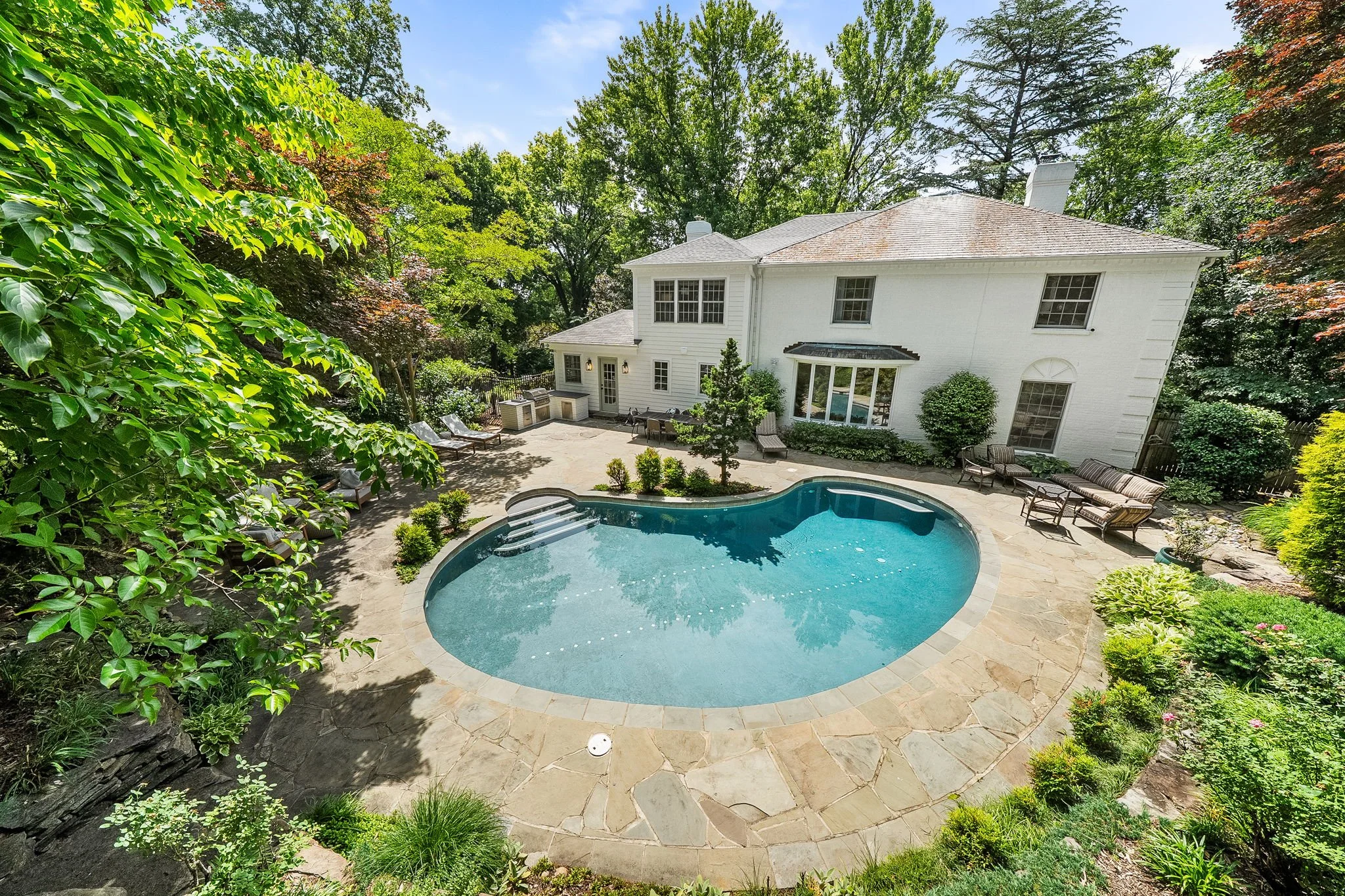 A backyard with a kidney-shaped swimming pool surrounded by a stone patio, lounge chairs, and lush green trees and bushes.