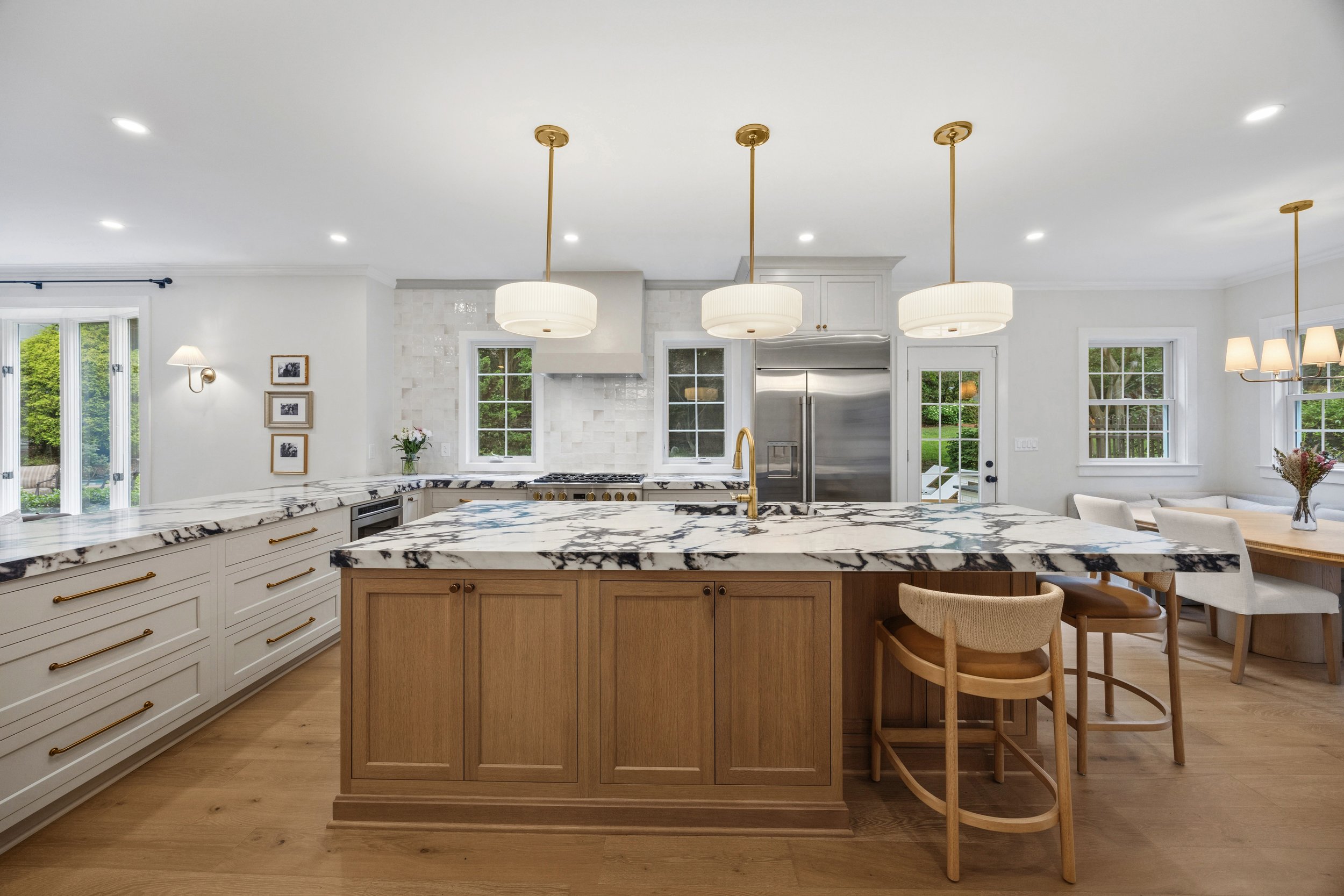 Modern kitchen with white cabinets, marble countertops, and gold pendant lights above island, large windows with green outdoor view.
