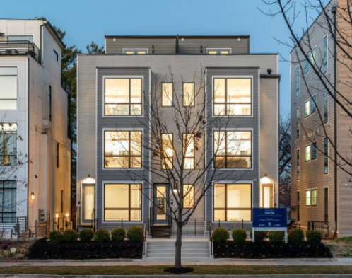 A modern, multi-story apartment building with large windows, a central tree in front, and a neatly landscaped sidewalk, during early evening.