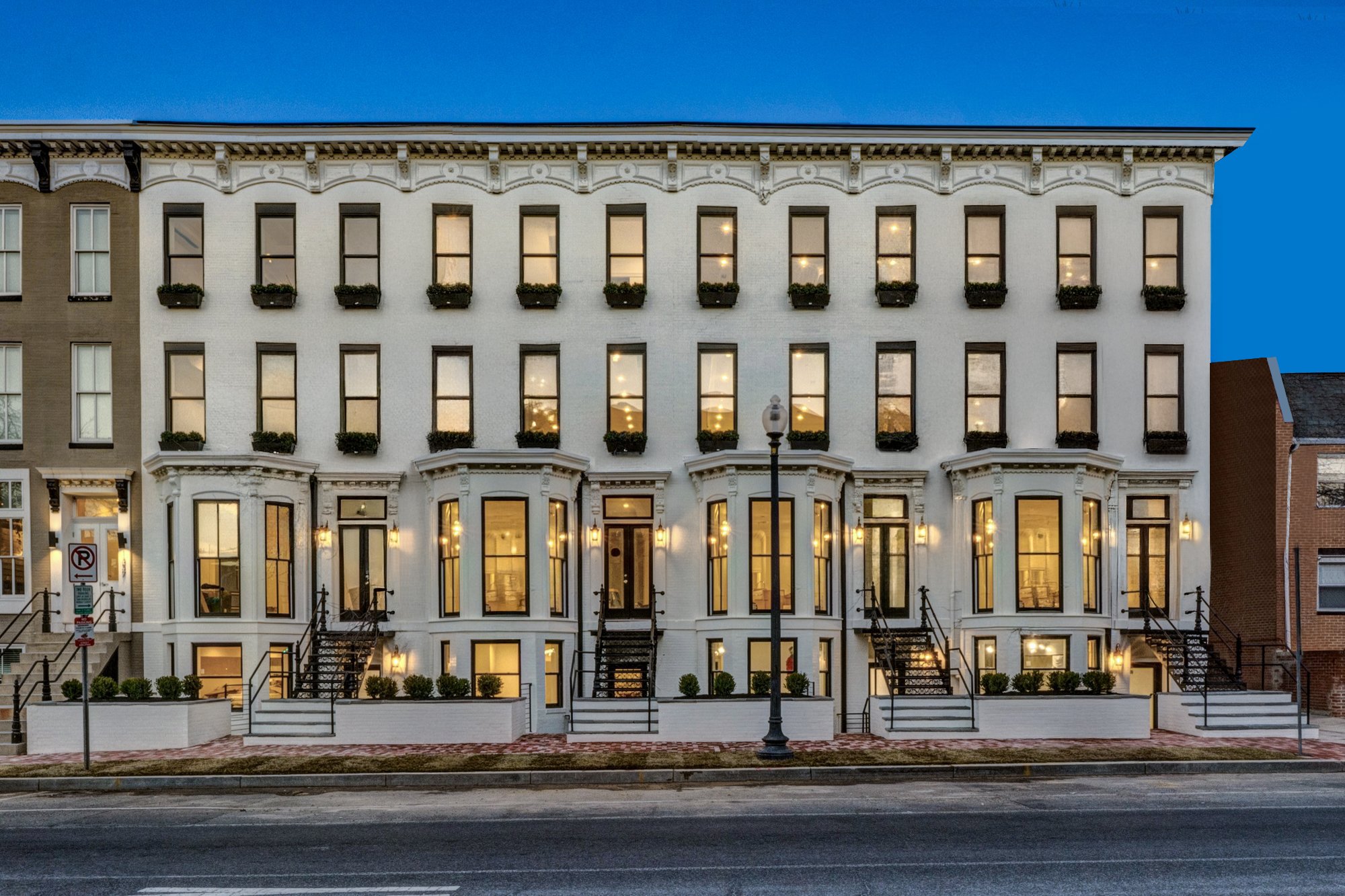 The image shows a three-story building with a white exterior, large ground-floor windows, and multiple rows of smaller upper-floor windows with flower boxes. The building is illuminated from inside, with lights visible through the windows. In front, there is a sidewalk, a street with lane markings, street lamps, and bike racks. The sky above is clear, indicating early evening or dusk.