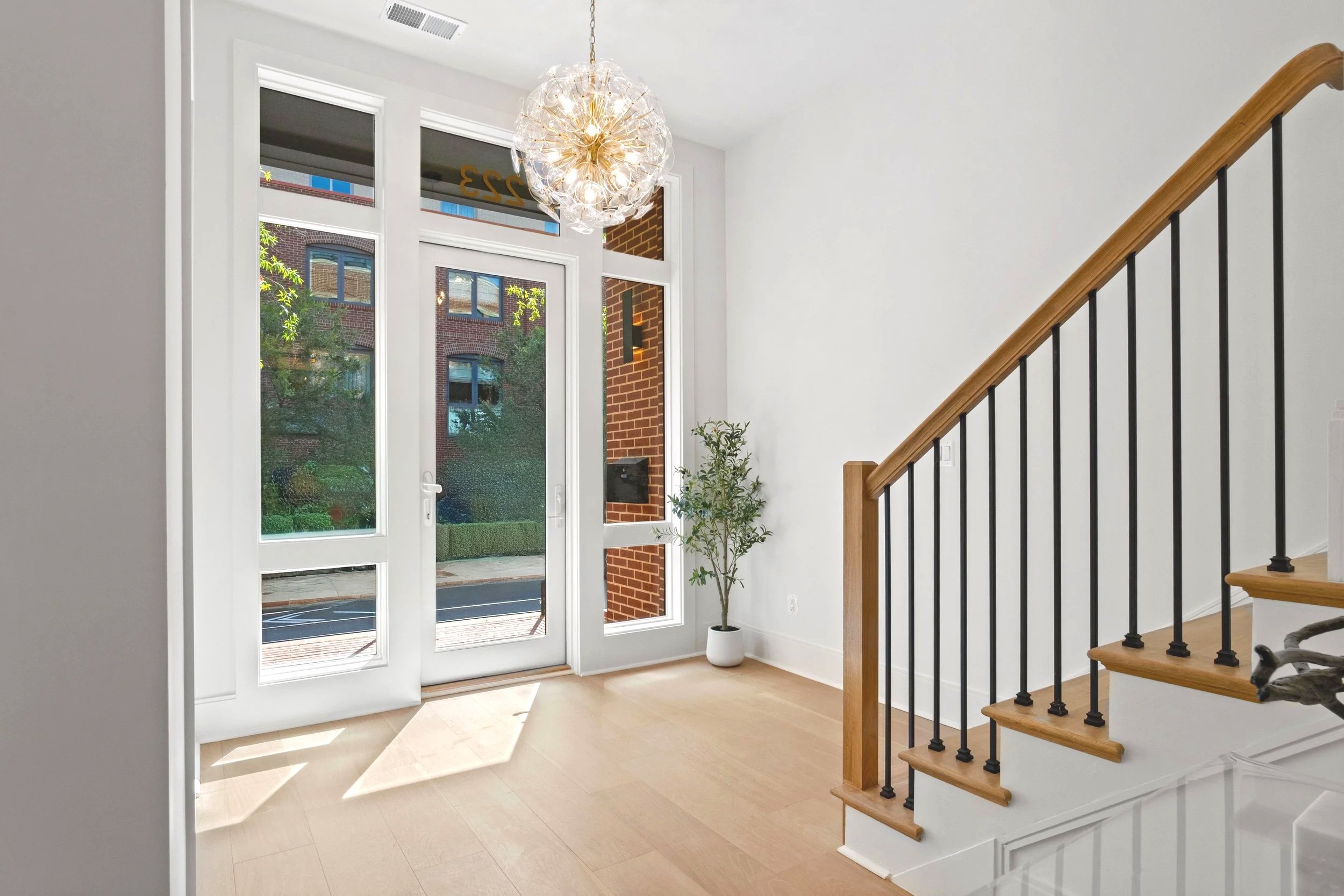Interior view of a modern house entryway with a glass front door, large windows, a stairway with wooden handrail and black balusters, a potted plant, and a decorative ceiling light fixture.