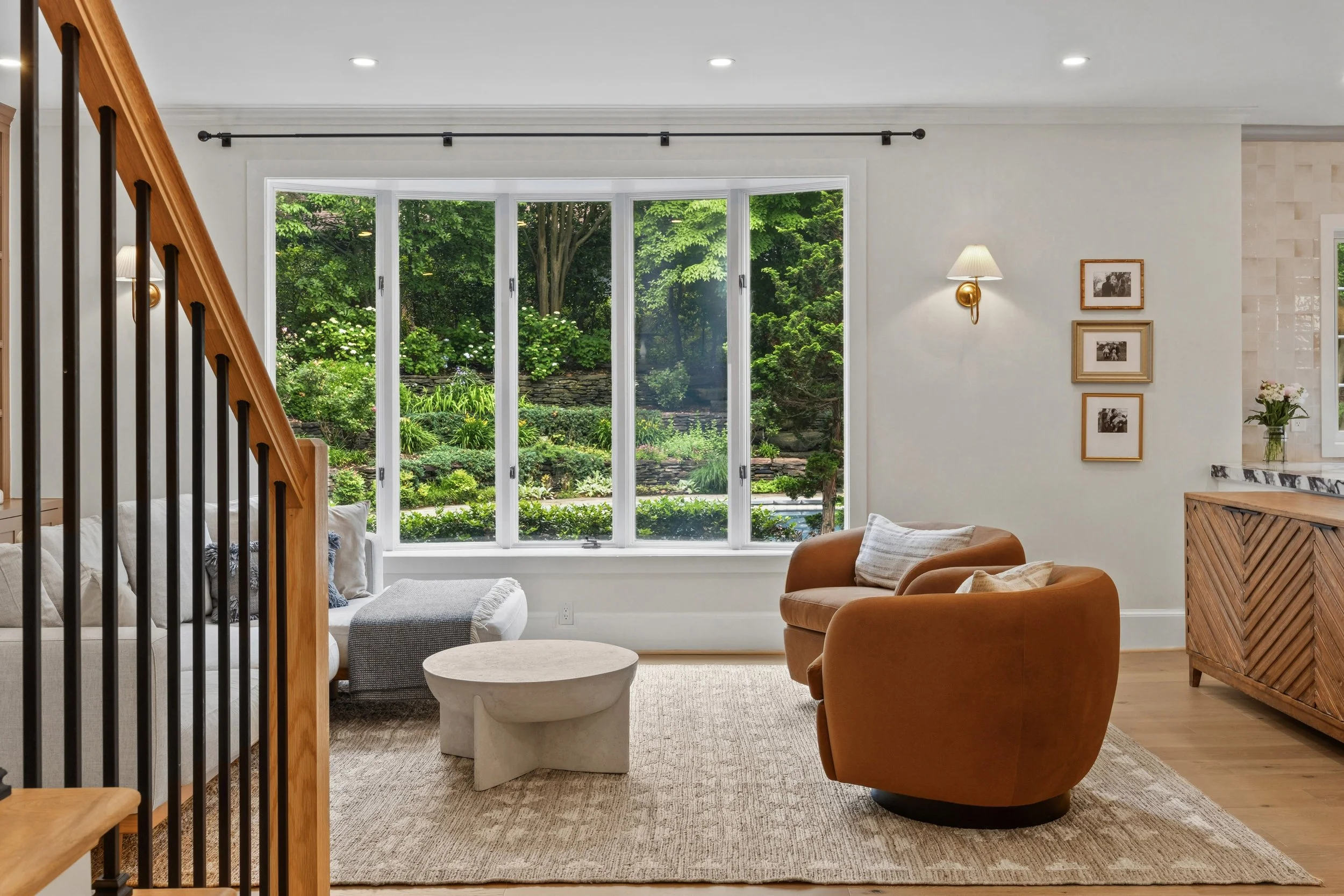 Living room with large bay window overlooking a lush green garden. Furnished with a white sofa, an orange armchair, a beige ottoman, and a wooden cabinet. Wall-mounted lamp and framed photos on the wall.