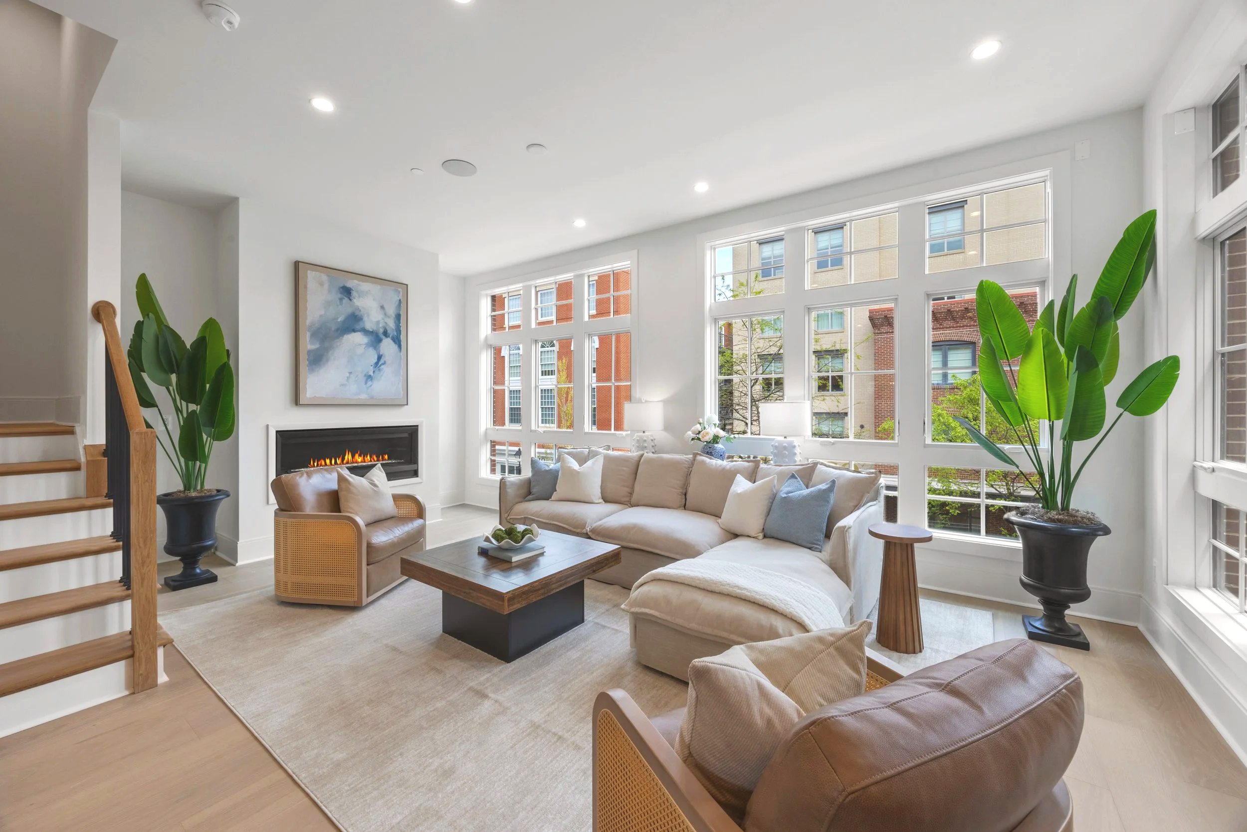 Bright living room with large windows, white walls, tan leather sofa and armchairs, modern fireplace, green plants, abstract wall art, coffee table, and natural light.