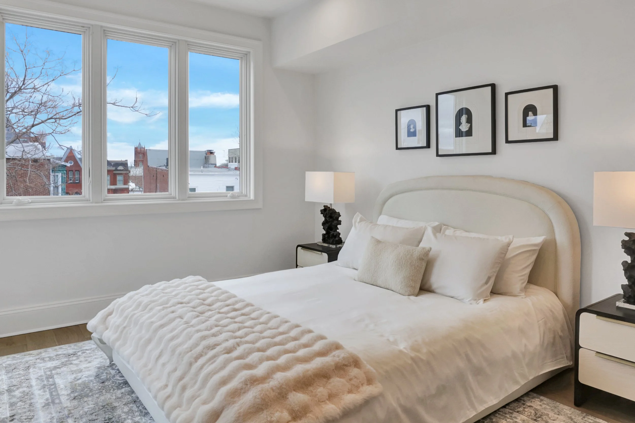 A bedroom with a large bed covered in a plush, beige comforter and multiple pillows, beside a nightstand with a black sculpture-shaped lamp. The wall above the bed has three framed pictures, and there are three large windows showing an urban cityscape with buildings and a cloudy sky.