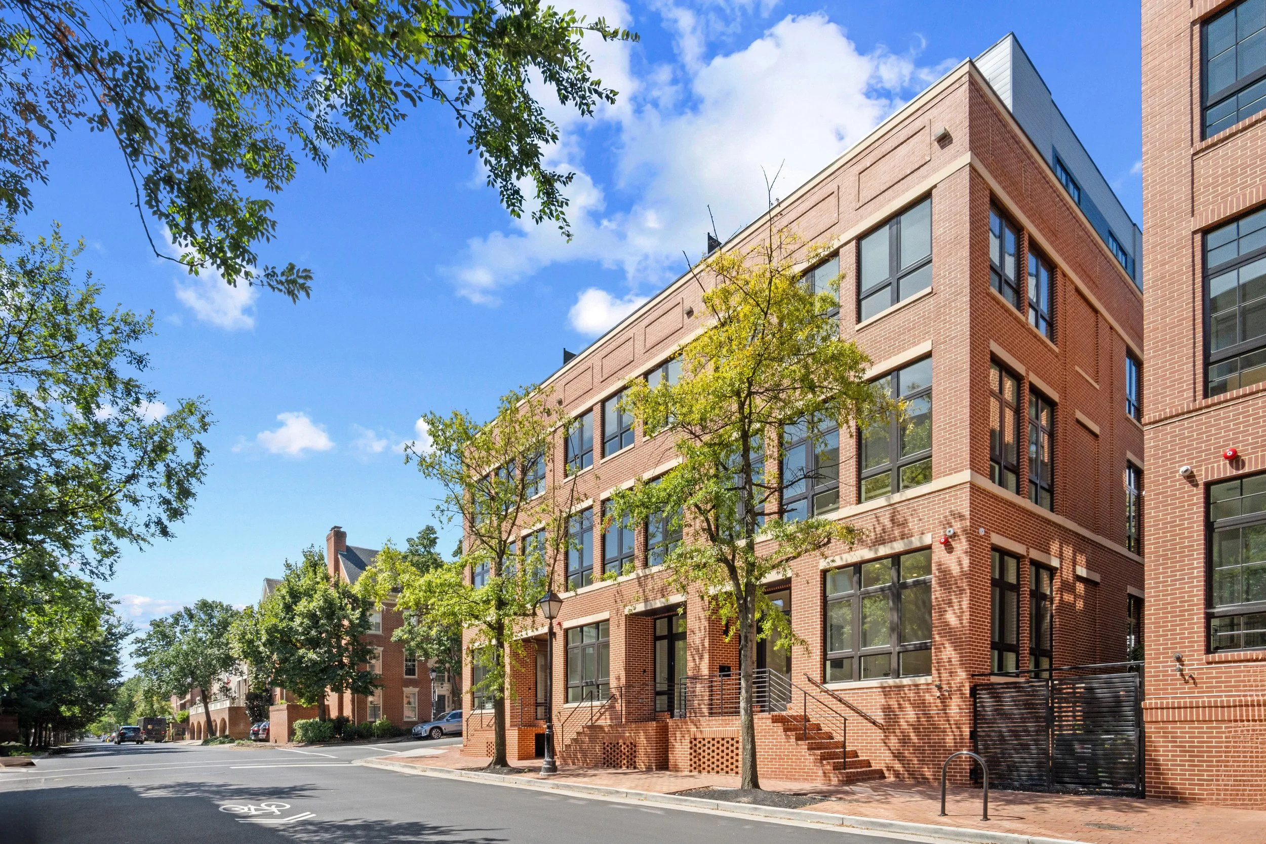 A multi-story brick apartment building on a city street with trees and parked cars, under a blue sky with clouds.