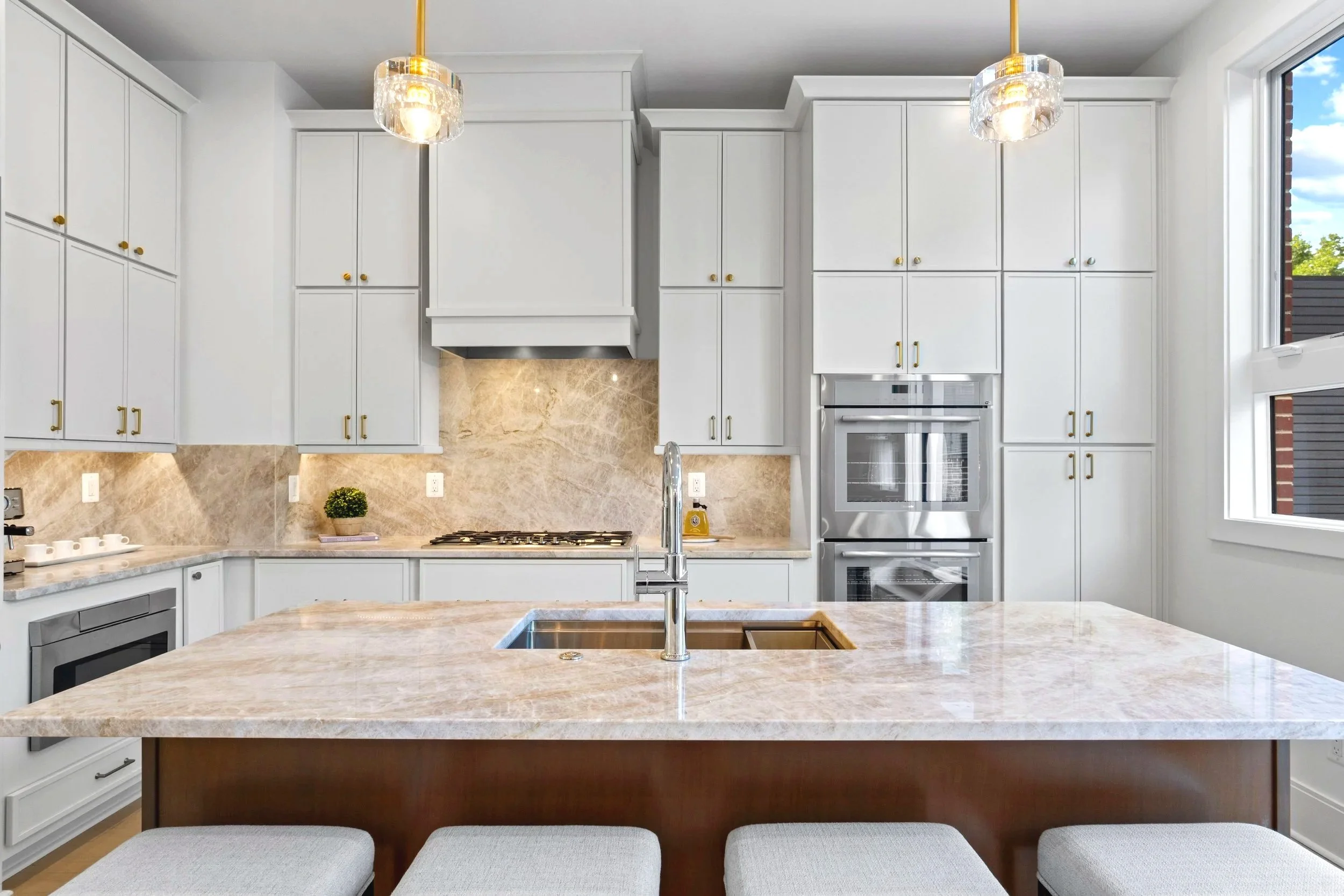 Modern kitchen with white cabinets, marble countertops, stainless steel appliances, pendant lights, and large window showing outdoor scenery.