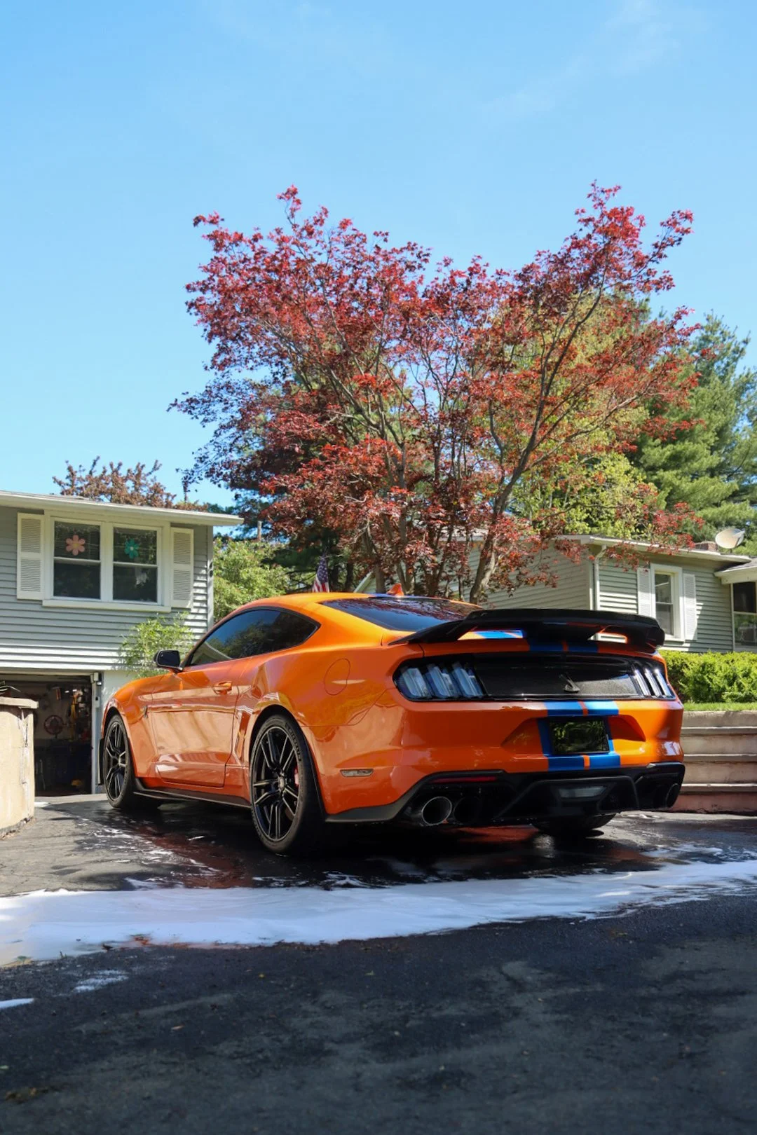 Orange sports car with blue racing stripes parked in driveway with soap and water on the ground, large tree with red leaves in background, and a house with gray siding and white windows.