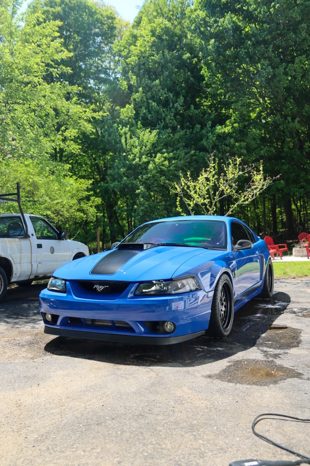 A blue Ford Mustang sports car with black racing stripes parked on a wet driveway during daytime. In the background, there's a wooded area with green trees, a white vintage truck, and red chairs.