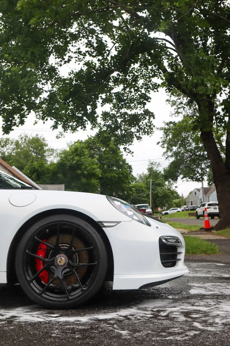 Close-up of a white sports car with black wheels and red brake calipers parked on a wet road with trees and houses in the background.