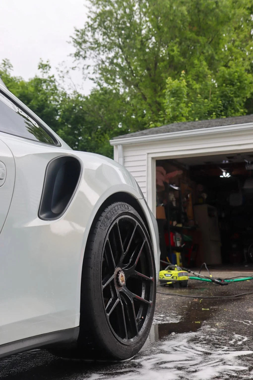 Close-up of a white sports car with a black wheel, parked outside a garage with car washing equipment and soap suds on the ground, and green trees in the background.