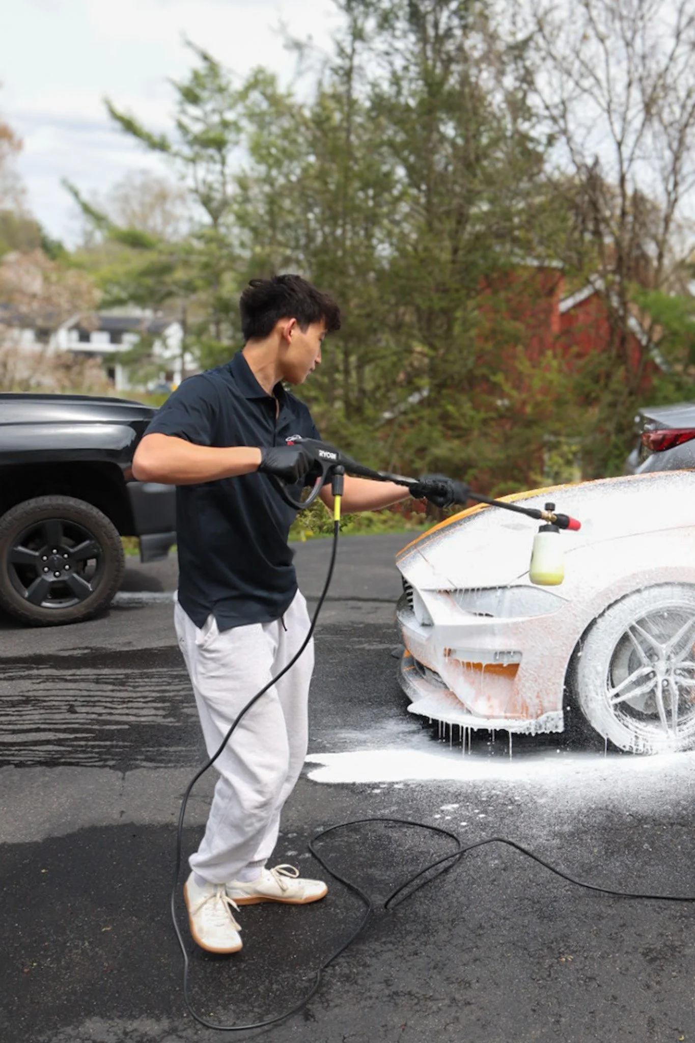 A man in a black shirt and white pants pressure washing a white sports car with foam. The car's front bumper and hood are covered in soap, and the man is holding a pressure washer wand. In the background, parked cars and trees with autumn foliage are visible.