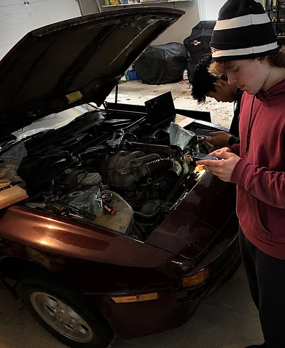 Two young men examining a car engine with the hood open, in a garage or workshop. One person in a red hoodie and black and white beanie is looking at their phone, while the other person is also focused on the engine. Various tools and equipment are around.