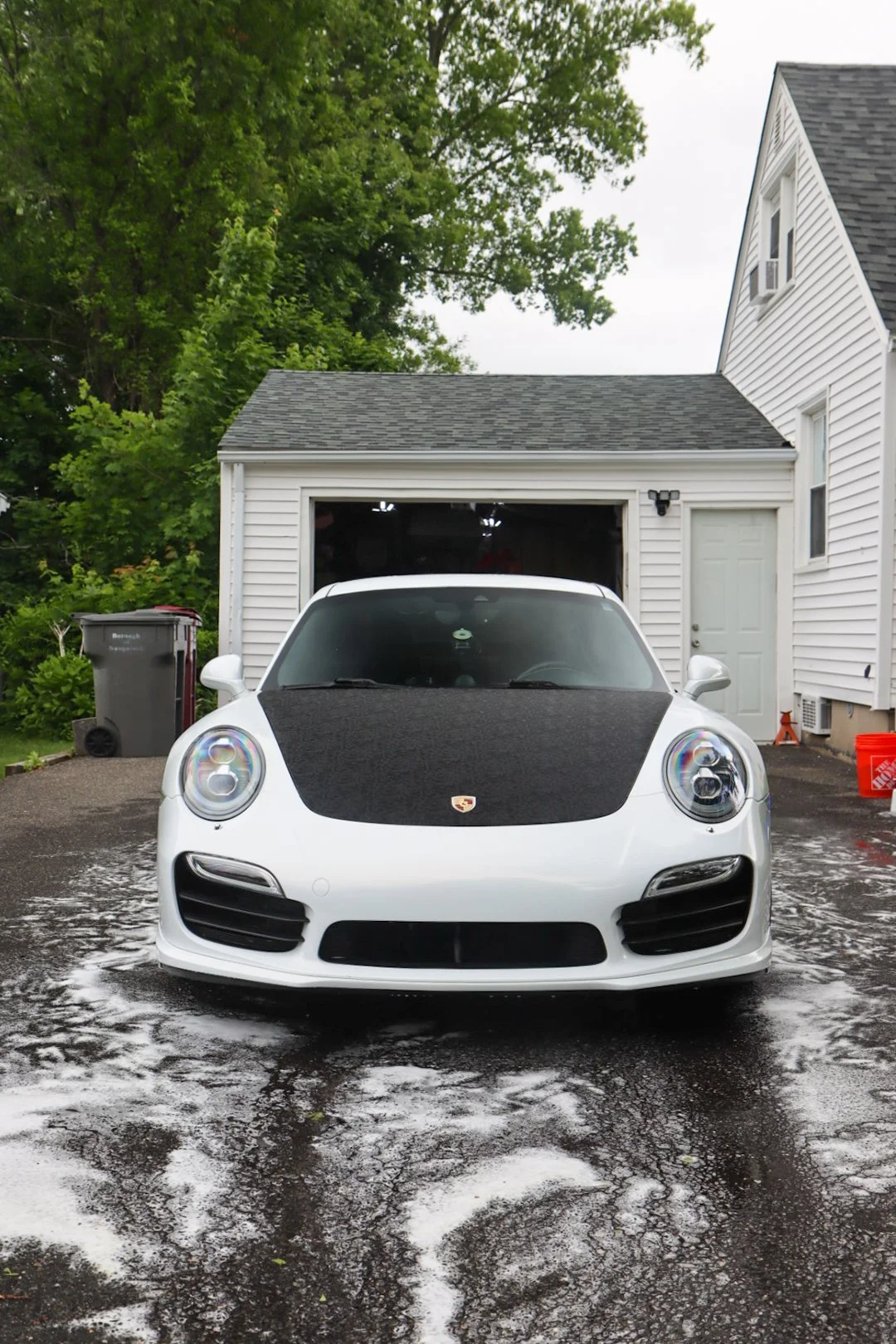 A white Porsche parked in a driveway with a wet surface and soap suds. There is a garage in the background with a partially open door, a trash bin, and a house with white siding and green trees nearby.