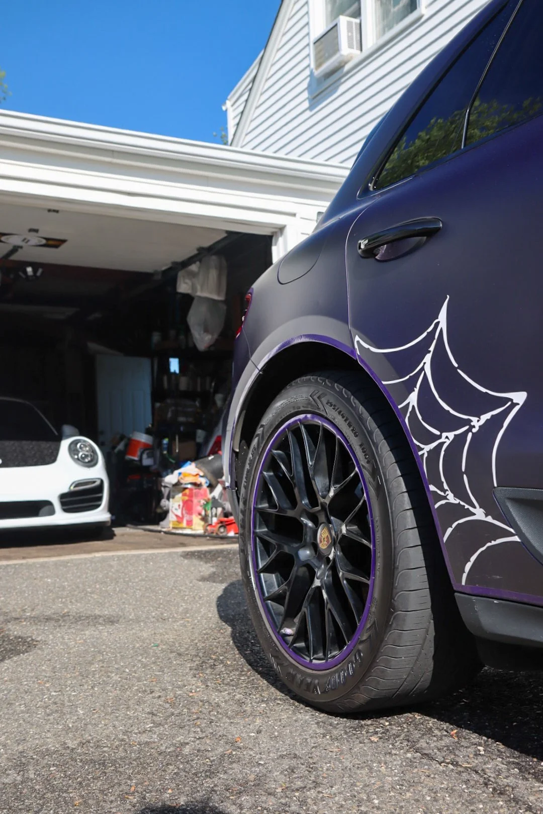 A black car with a white spider web decal on the side is parked in front of a garage. The garage door is open, revealing clutter inside, and another white car is parked inside the garage. The sky is clear and blue, and the house has white siding.
