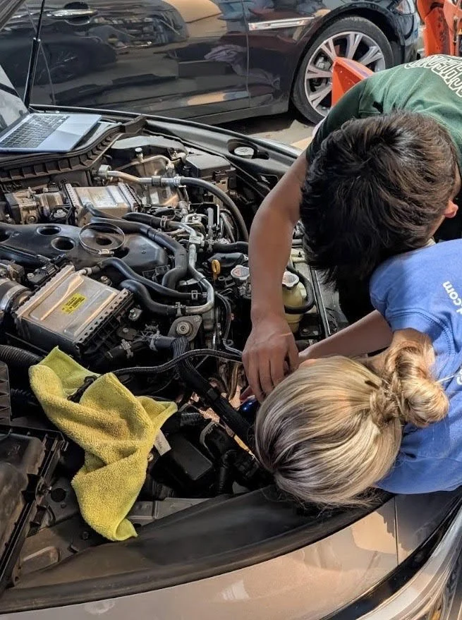 Two people working under the open hood of a car in a garage, inspecting the engine with a laptop nearby.