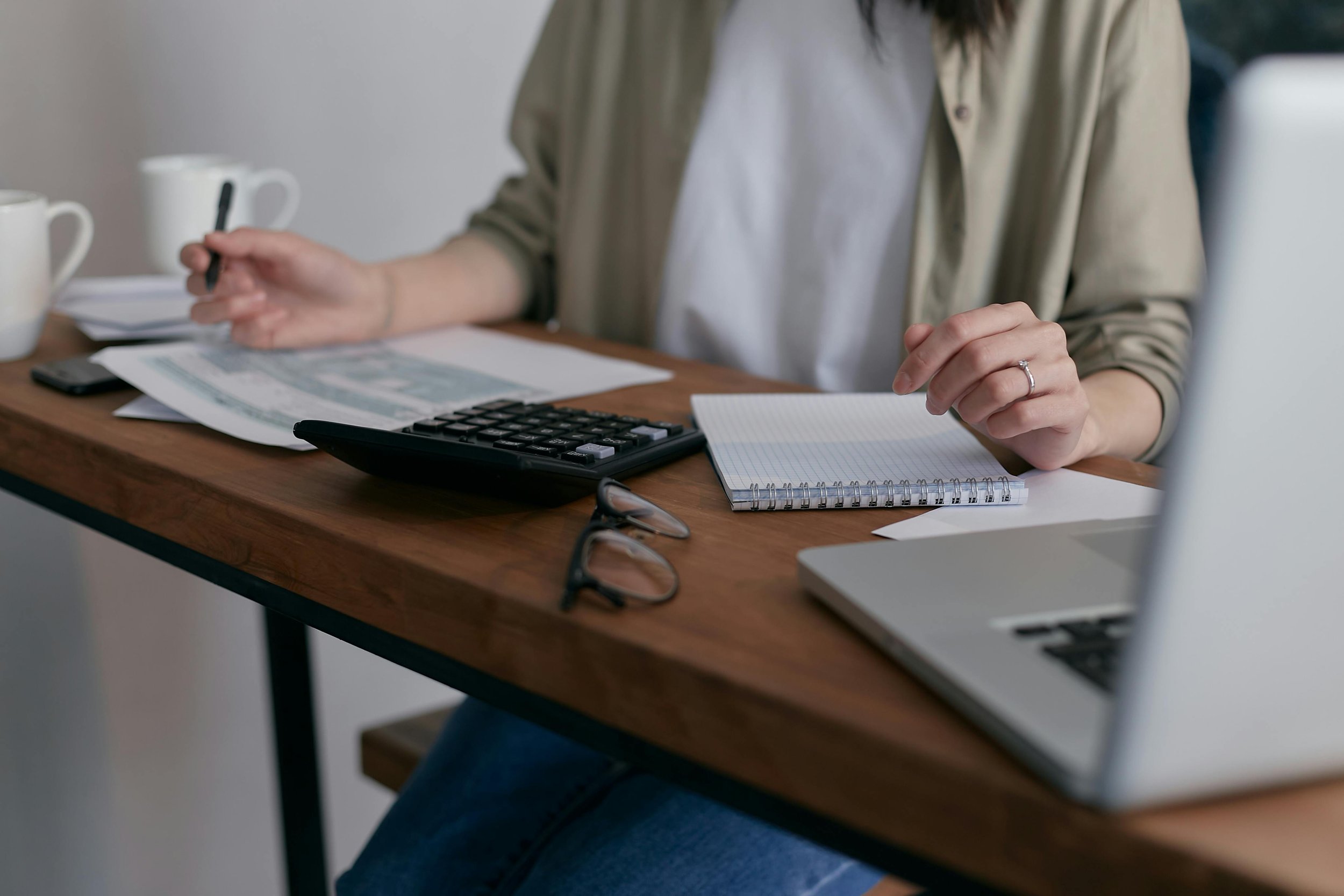 Person working at a wooden desk with notebooks, a calculator, glasses, a laptop, and coffee mugs.