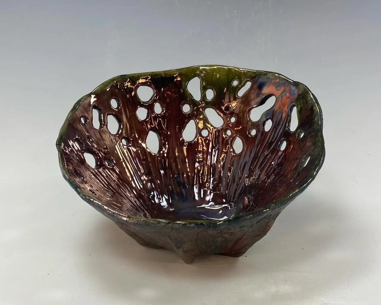 A small, irregularly shaped bowl made of glass or ceramic with a dark, iridescent, and marbled surface. The bowl features numerous holes of various sizes throughout its structure, resembling a net or lace pattern.