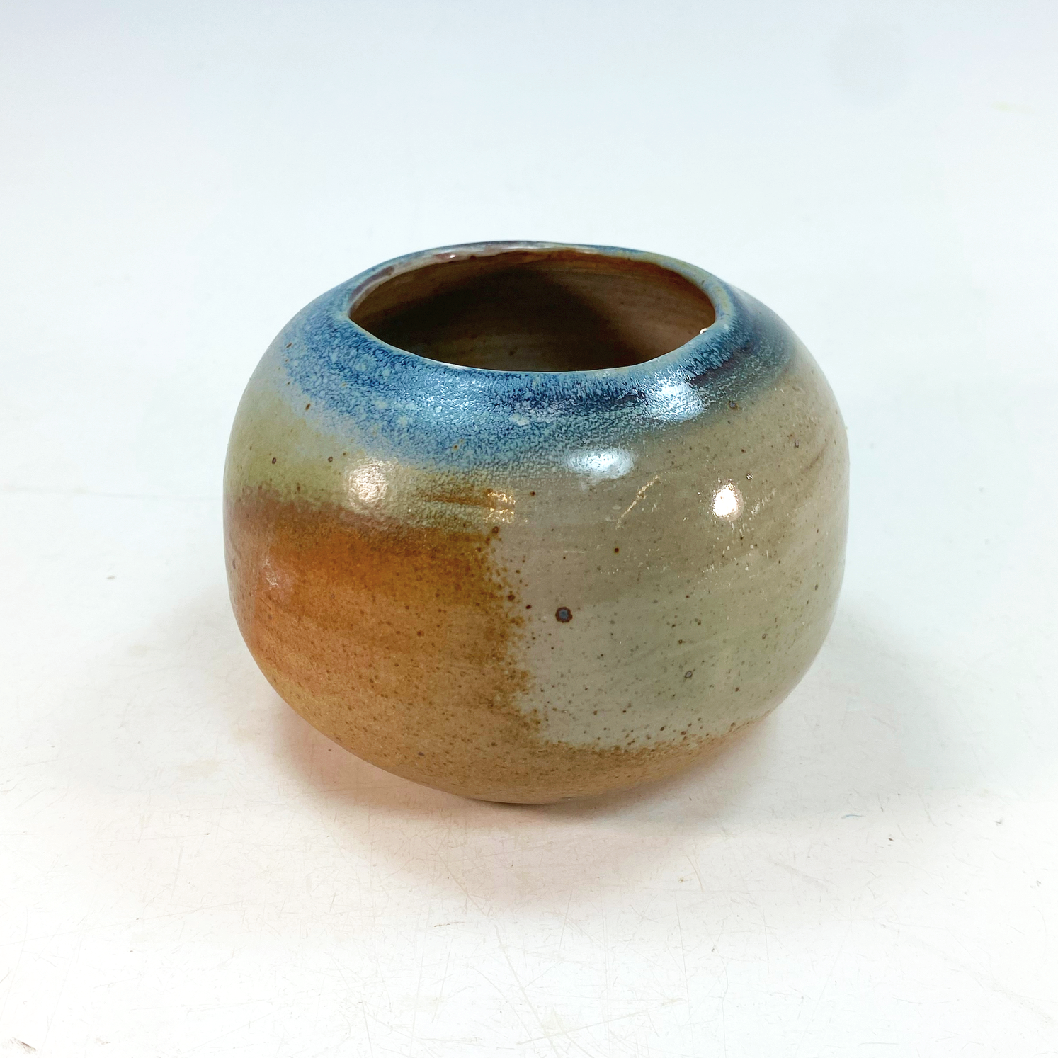 A round ceramic vase with a small opening at the top, glazed with a gradient of colors including beige, orange, blue, and green, placed on a white surface.