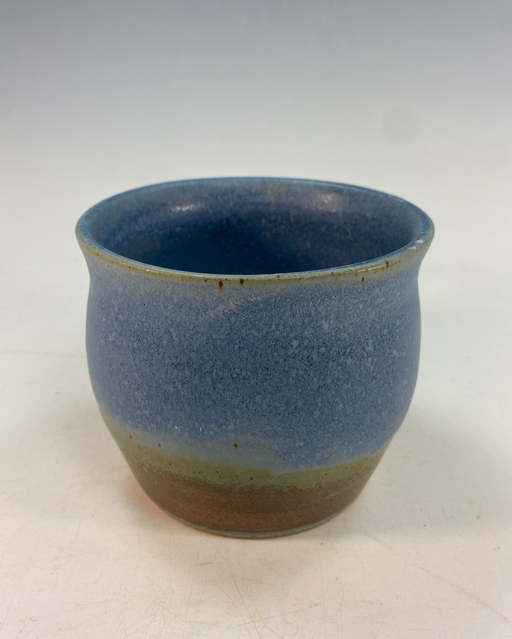 A ceramic cup with a blue glaze on the interior and a gradient glaze from green to brown on the outside, placed on a white surface.