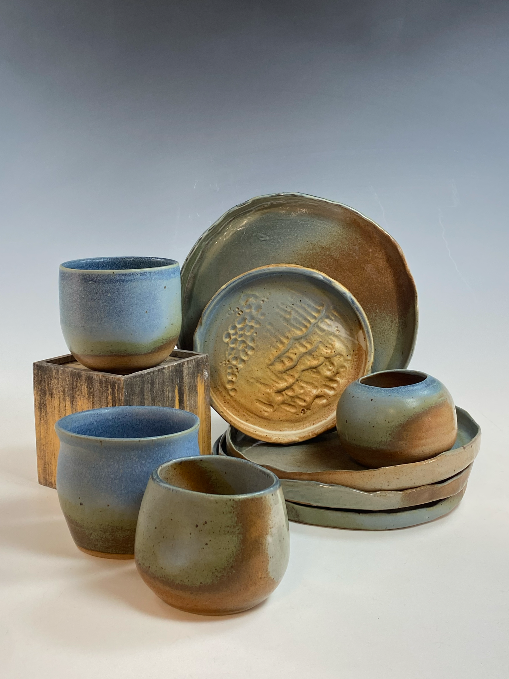 Collection of handmade pottery cups, bowls, and plates with earthy tones and textured glaze, displayed on a wooden box and a white surface against a plain background.