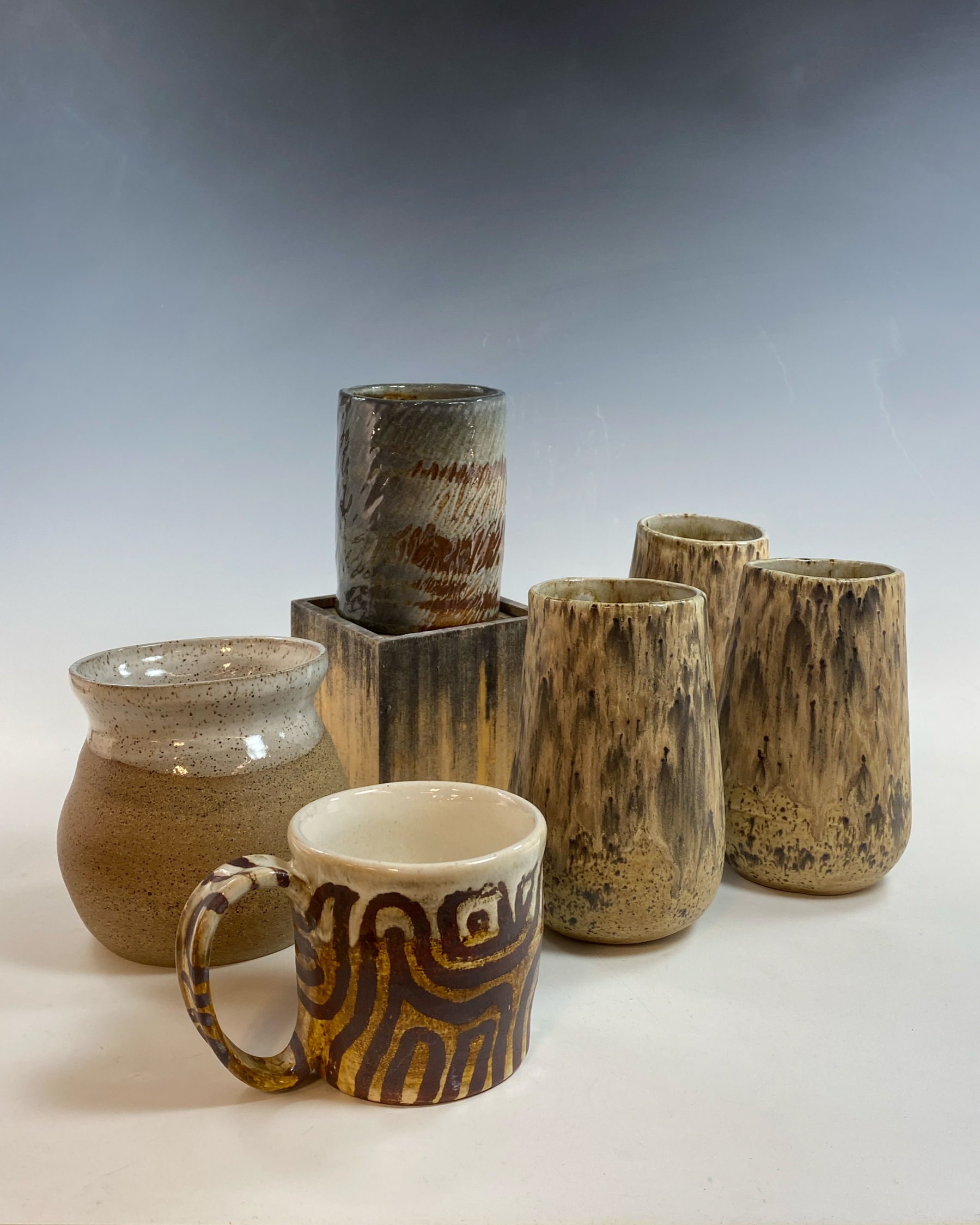A collection of ceramic cups and vases with earthy tones and various patterns, arranged on a white surface against a gradient background.