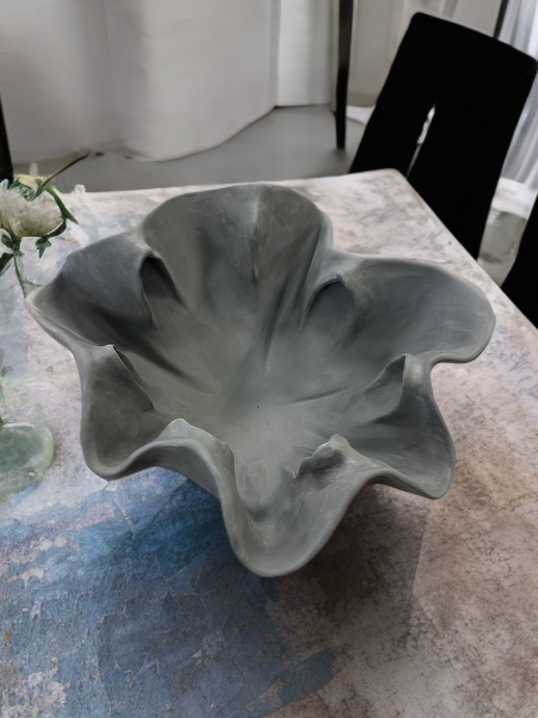 A gray, abstract, flower-shaped ceramic or porcelain bowl with wavy, petal-like edges, placed on a table with a textured tablecloth along with a small glass vase containing flowers.