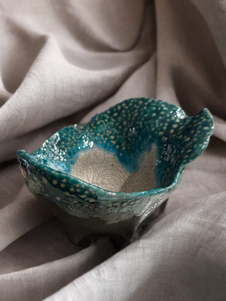 A decorative ceramic bowl with a textured, irregular rim, glazed in shades of teal and blue with a speckled pattern, placed on light-colored fabric.