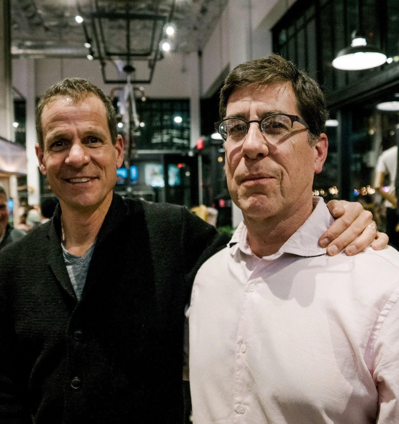 Two men posing for a photo inside a restaurant or café at night, with the man on the left smiling and the man on the right looking serious, with a hand on his shoulder.