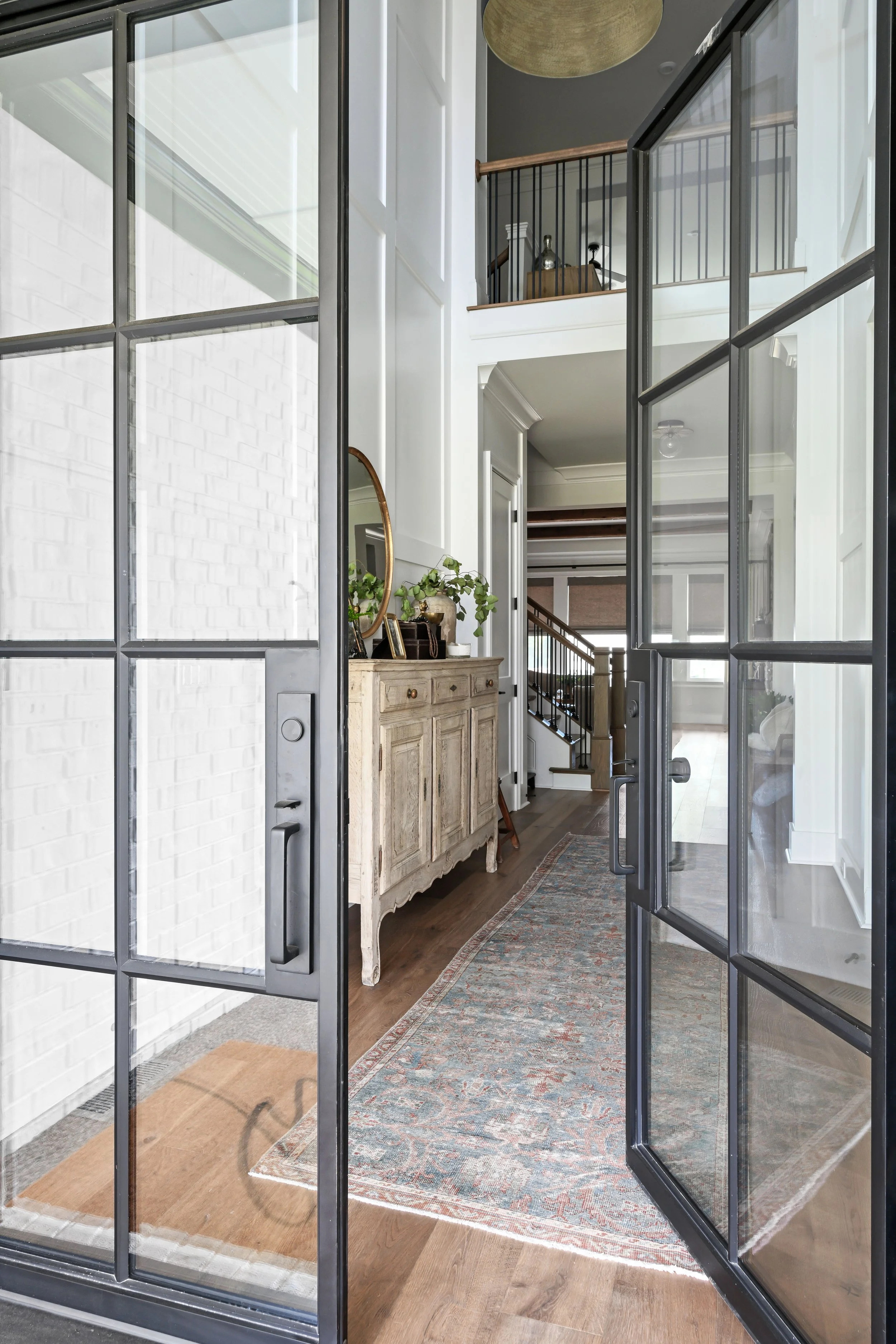 View through glass-paned black metal door into a bright home entryway with a textured white brick wall, a vintage wood sideboard with plants and decorative items, and a staircase with black metal spindles.