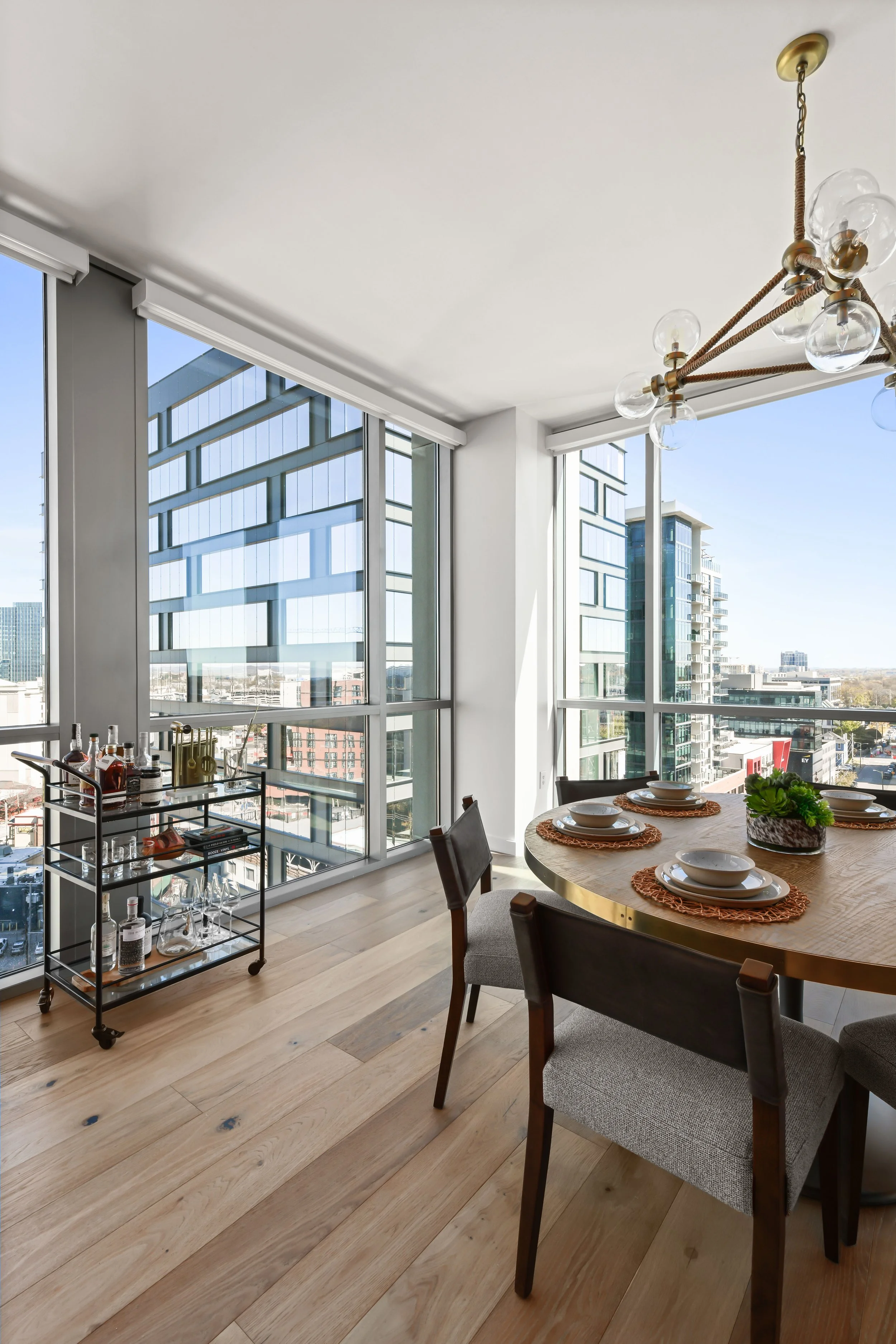 Modern dining area with large windows, round wooden table set with four plates and placemats, bar cart with bottles and glasses, and a contemporary chandelier with glass globes.