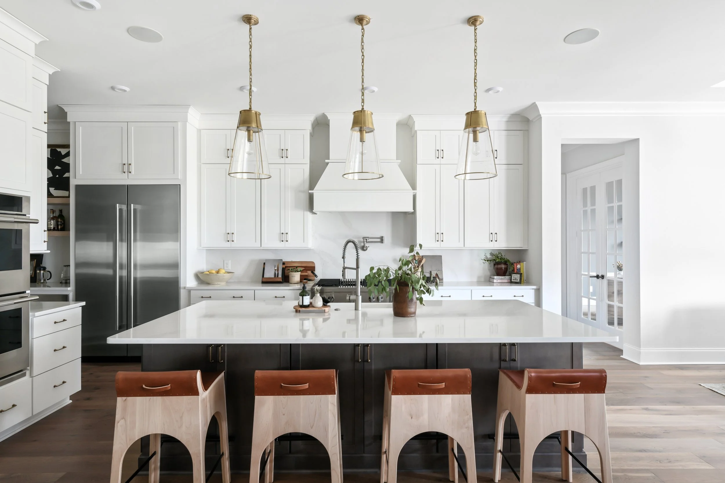 Modern kitchen with white cabinets, stainless steel appliances, a large island with a white countertop, and four wooden bar stools with brown leather seats. Hanging pendant lights above the island and hardwood flooring.