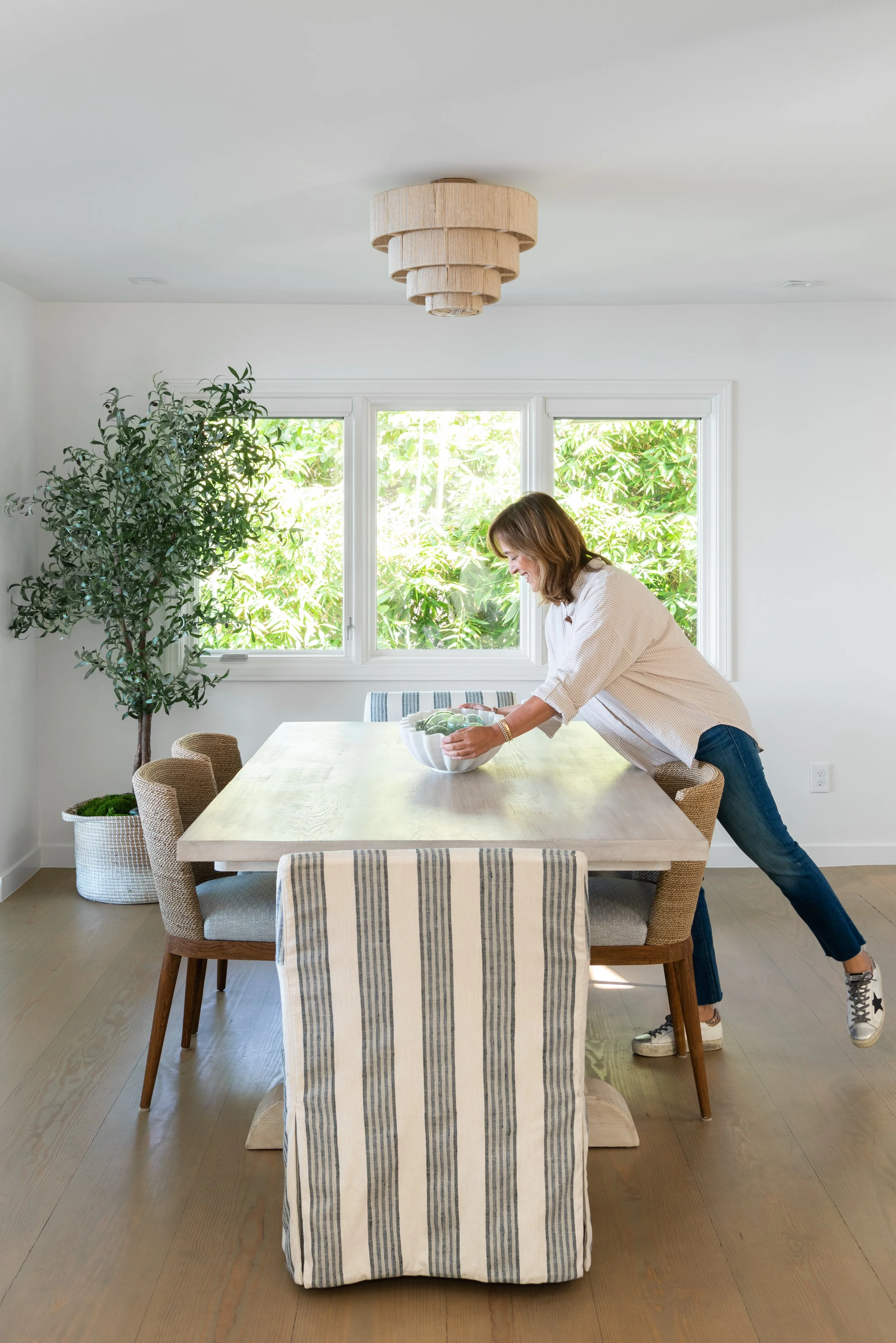 Woman leaning over a dining table, reaching for a green fruit in a bowl, in a bright dining room with a large window and indoor plant.