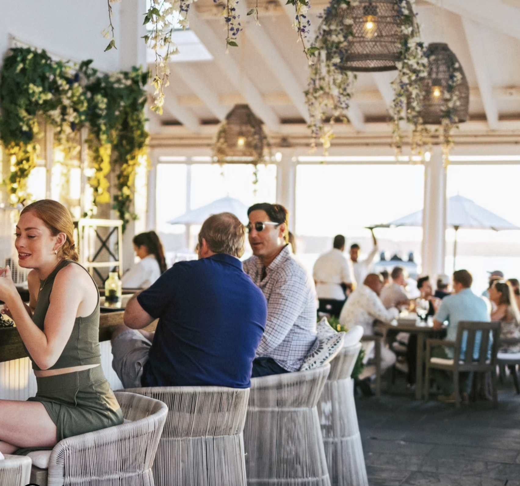 People dining at a restaurant with large windows, hanging light fixtures, and greenery decoration.
