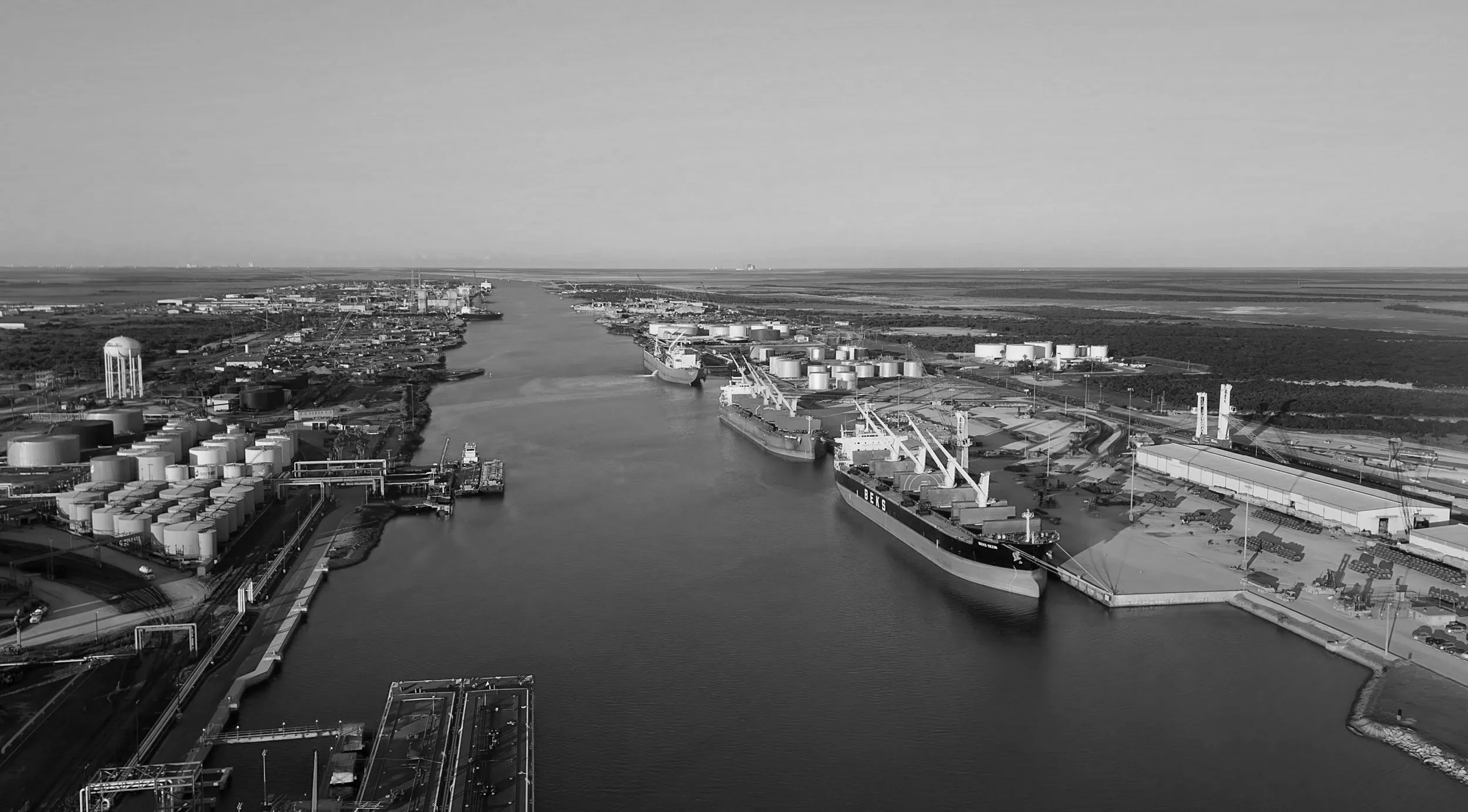Aerial view of an industrial shipping port with cargo ships, storage tanks, and cranes along a waterway in a flat landscape.