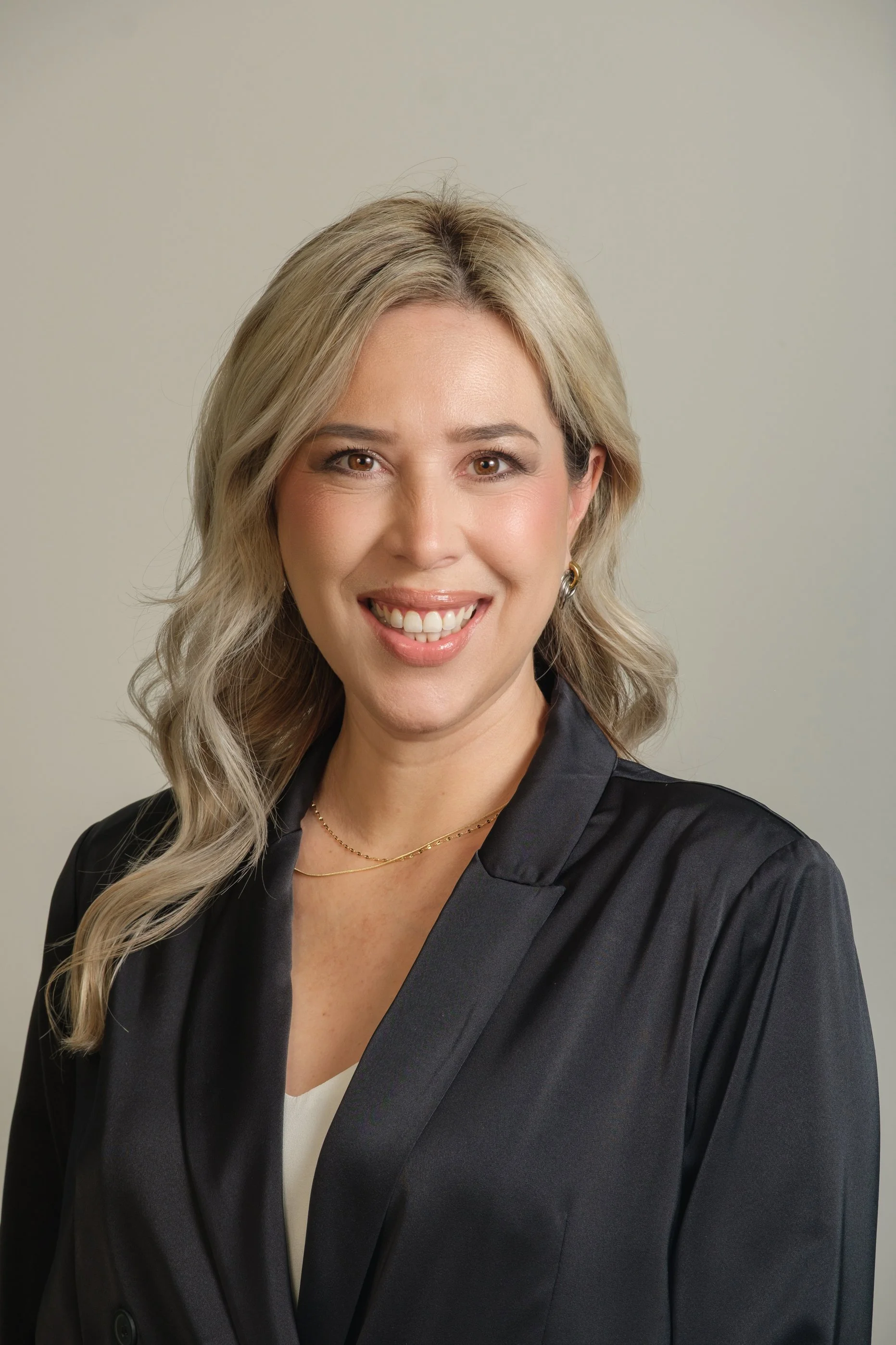 A woman with blonde wavy hair smiling, wearing a black blazer and gold jewelry, against a plain light background.
