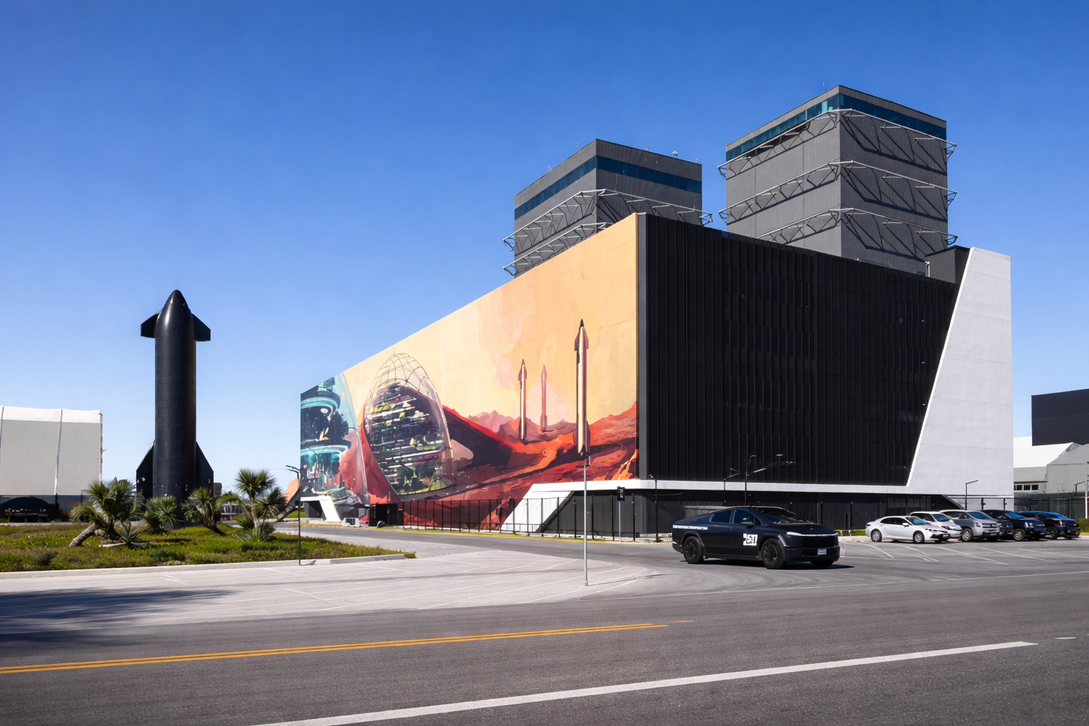 Modern building with a large space-themed mural, a rocket statue nearby, and a parking lot with several cars.