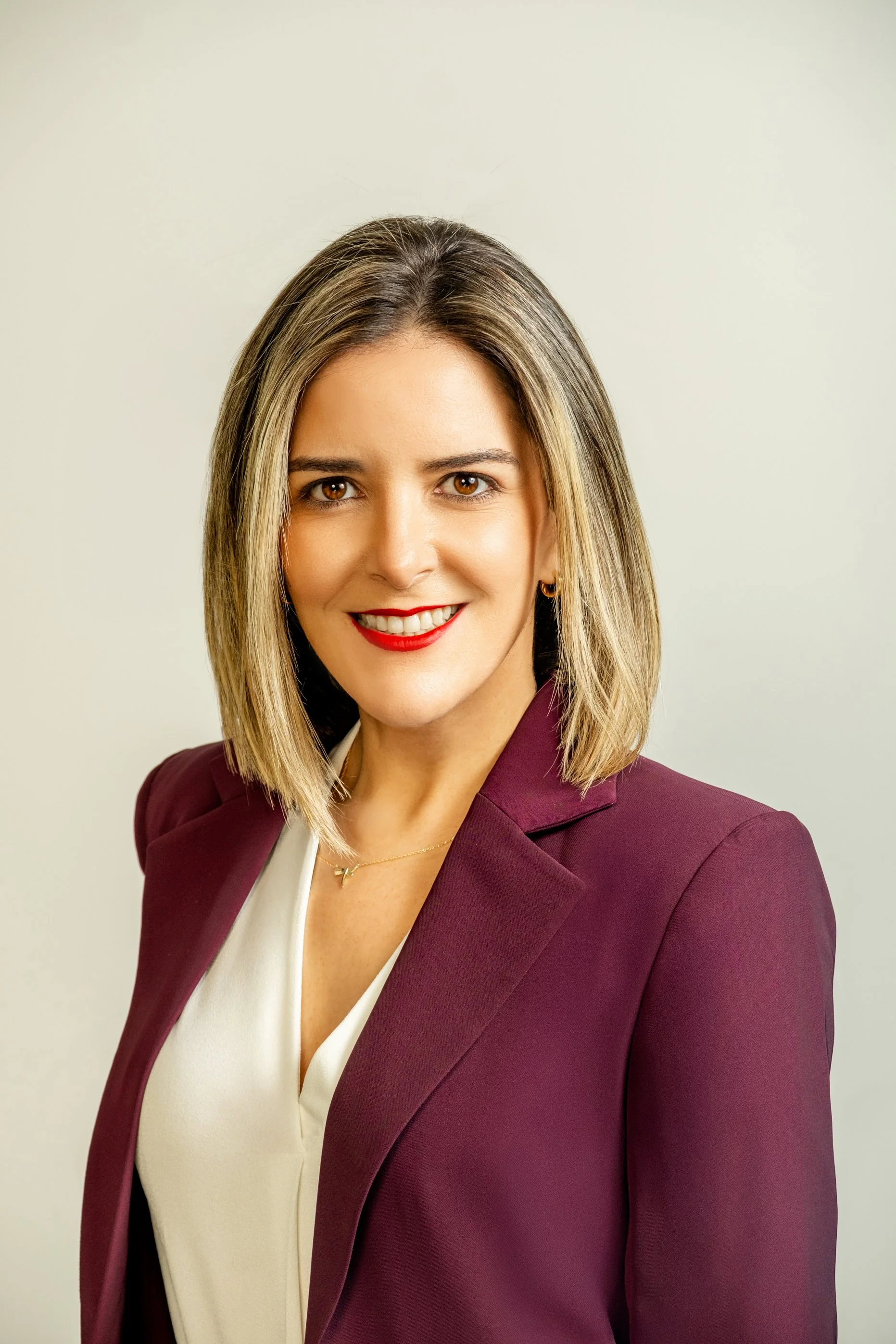 Professional woman with shoulder-length blonde hair, wearing a burgundy blazer and white blouse, smiling with red lipstick, against a plain light background.